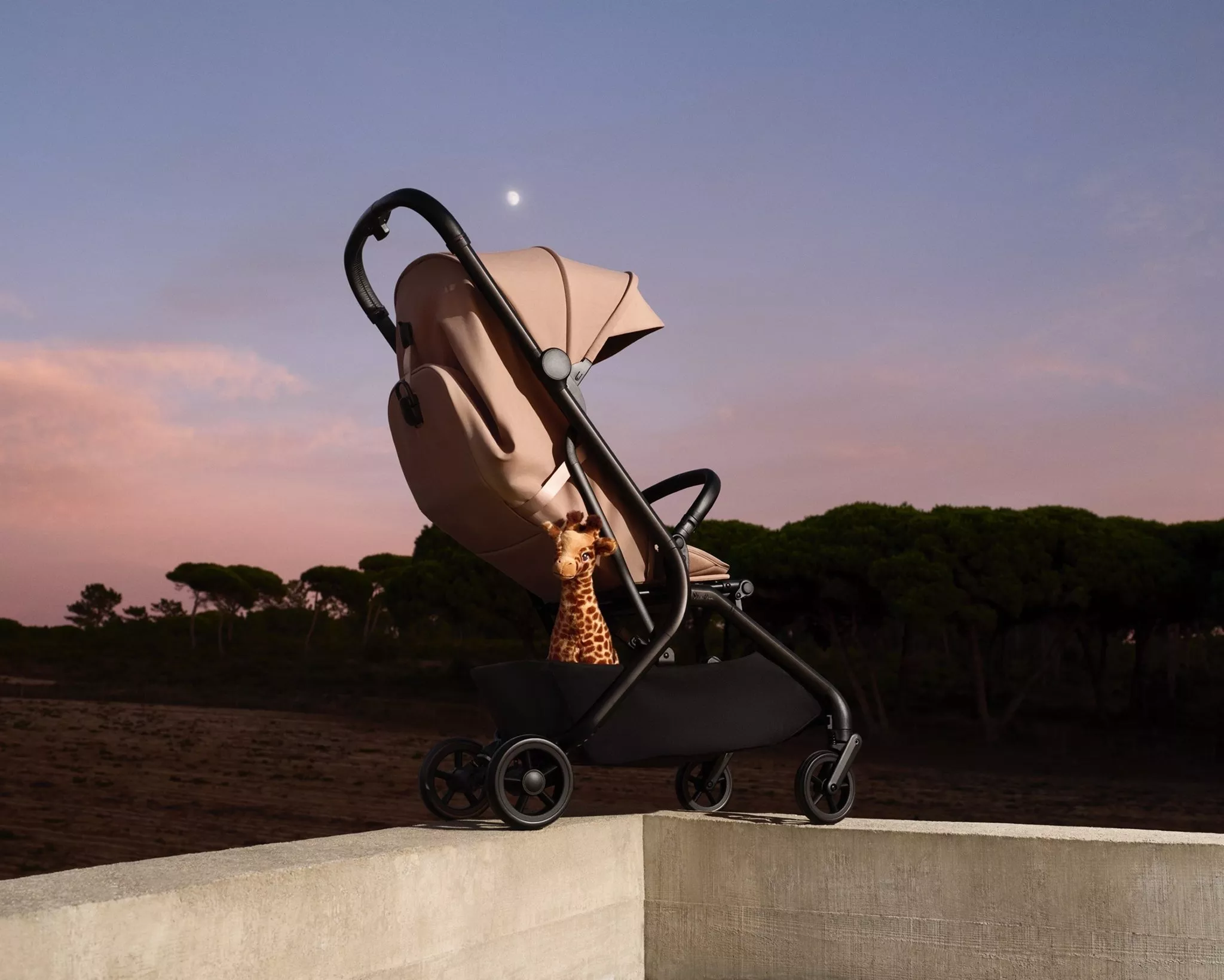Modern beige baby pushchair with toy giraffe, photographed outdoors at sunset with trees and moon in sky
