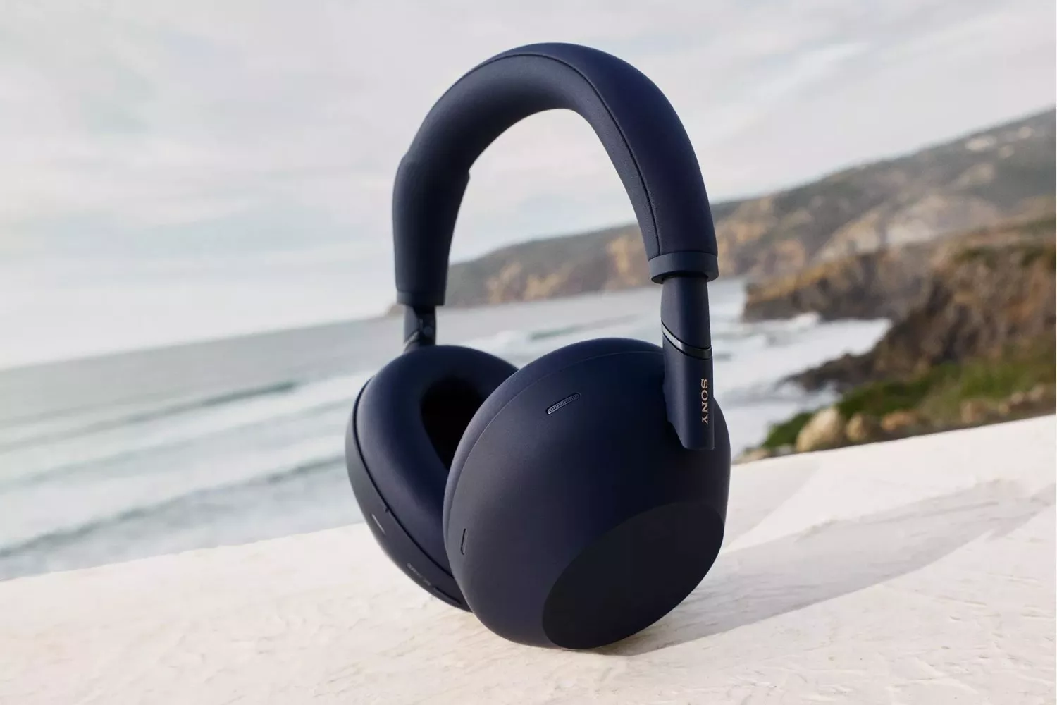 Sony over-ear noise-cancelling wireless headphones on a stone ledge with coastal sea and cliffs behind
