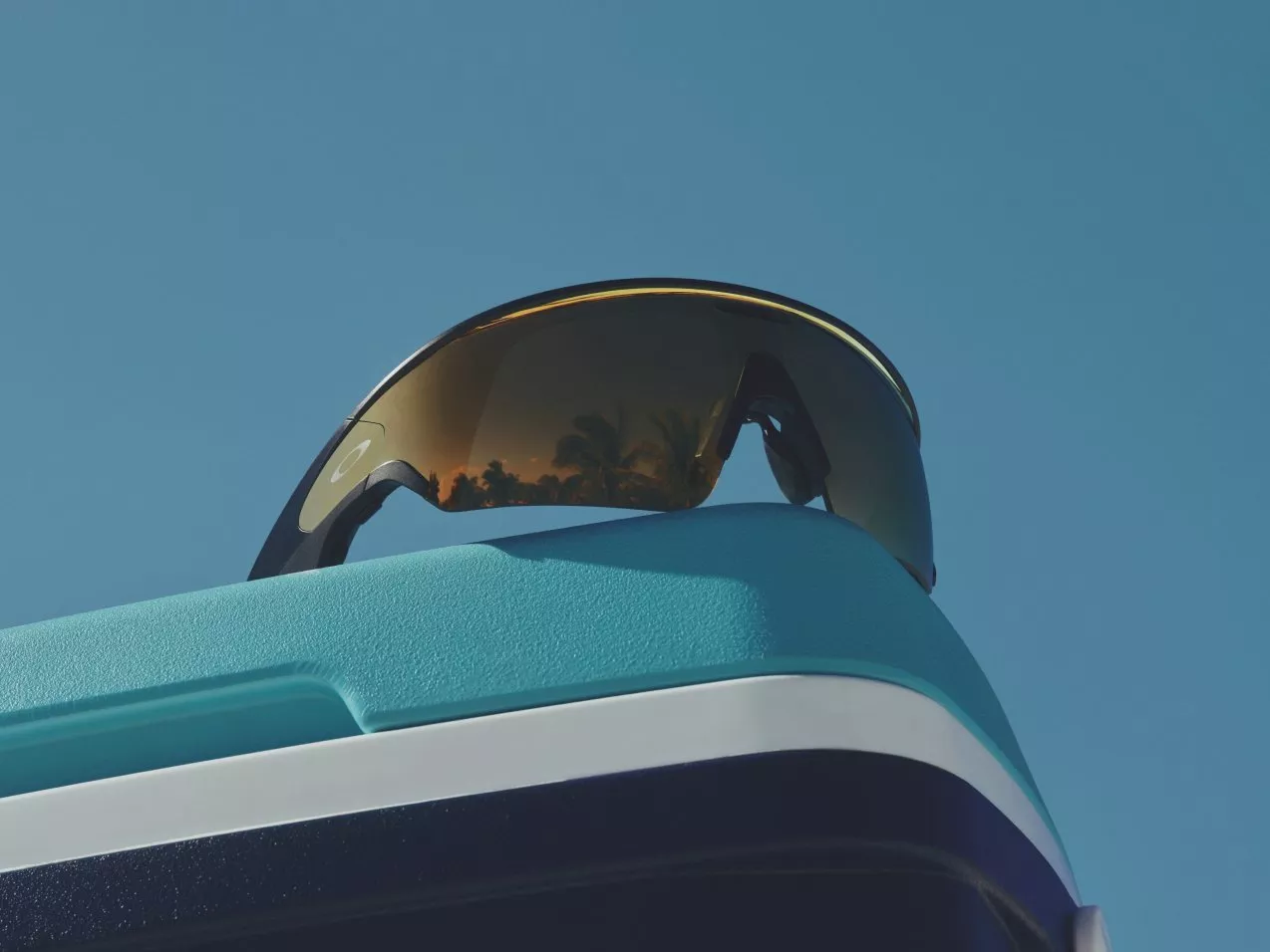 Polarised wraparound sunglasses resting on a teal cool box against clear blue sky, outdoor summer travel gear