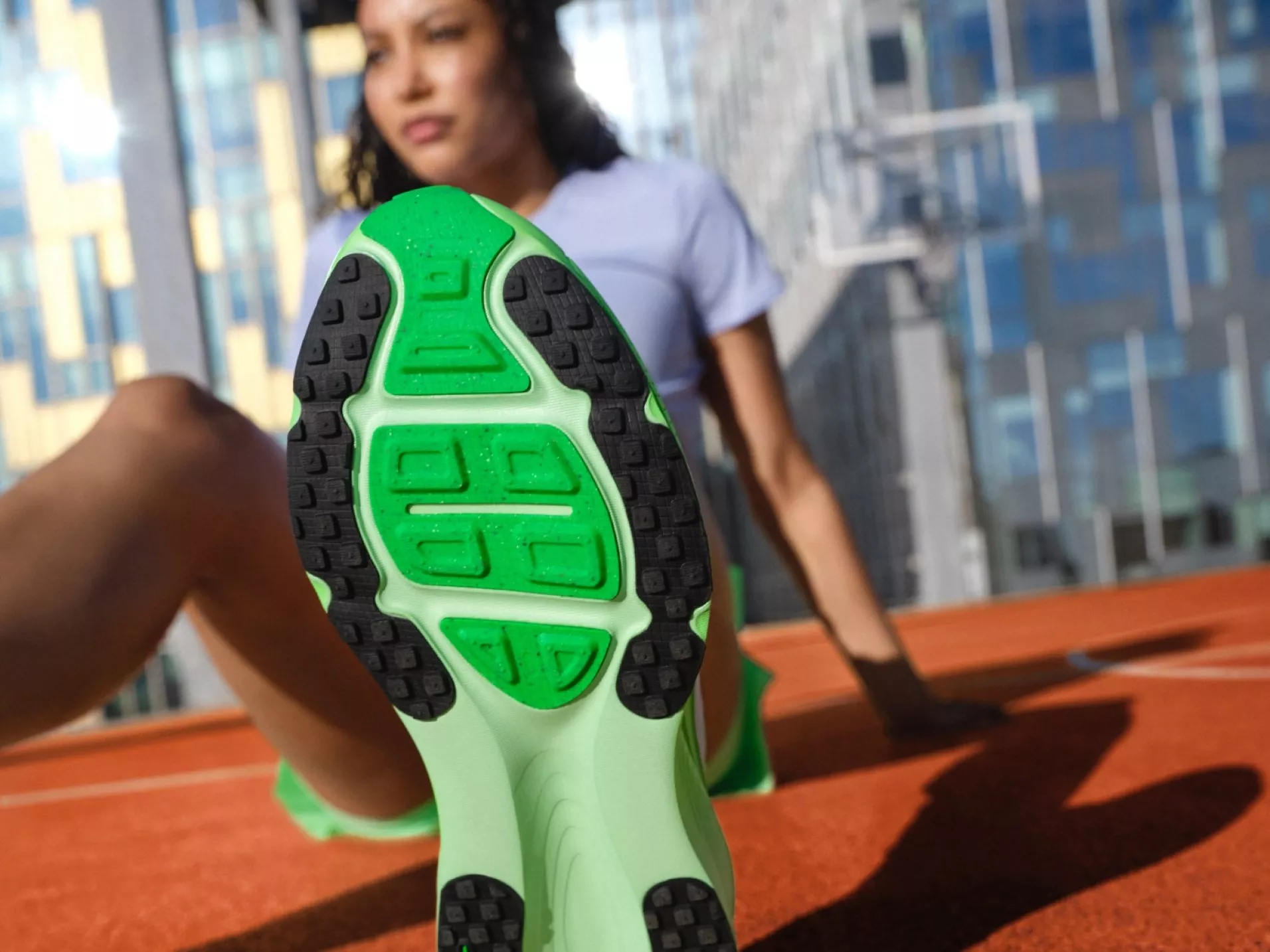 Close-up of green running shoe sole as woman stretches on outdoor track, city buildings in background