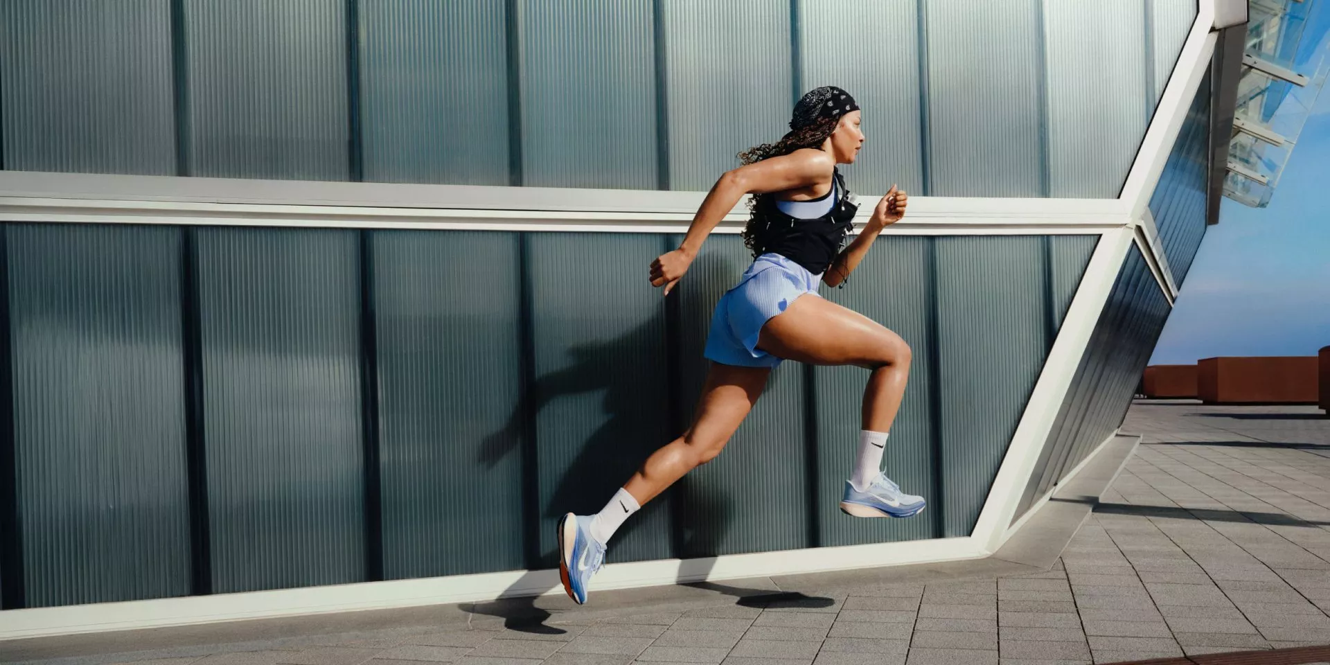 Female athlete sprinting outdoors by modern glass building, wearing running kit and trainers in daylight