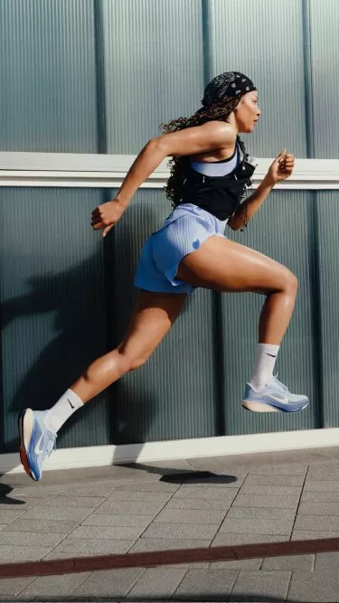 Female athlete sprinting outdoors in blue running kit and trainers, mid-stride beside modern building wall