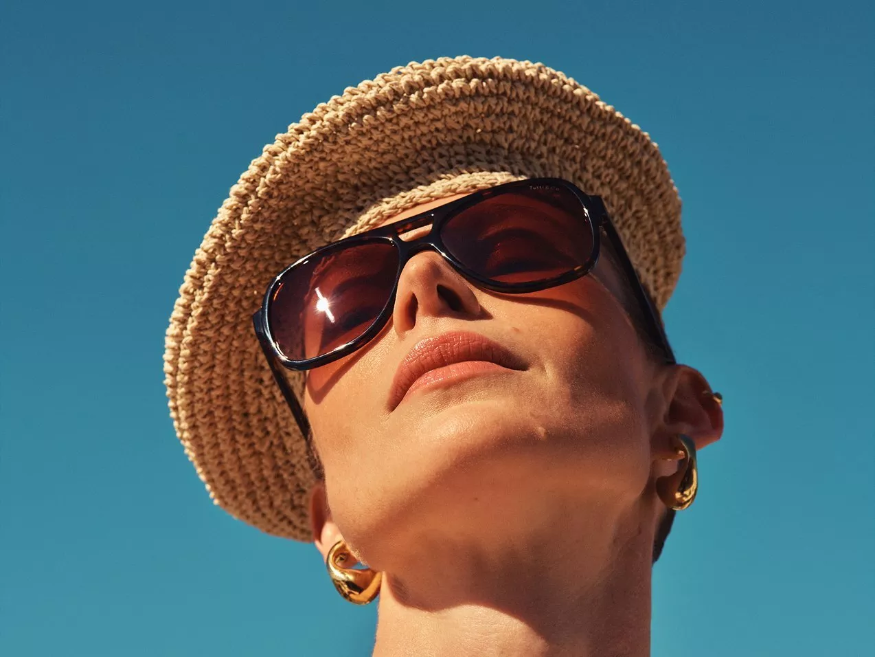 Close-up of person in woven straw sun hat, sunglasses and gold hoop earrings against blue sky background