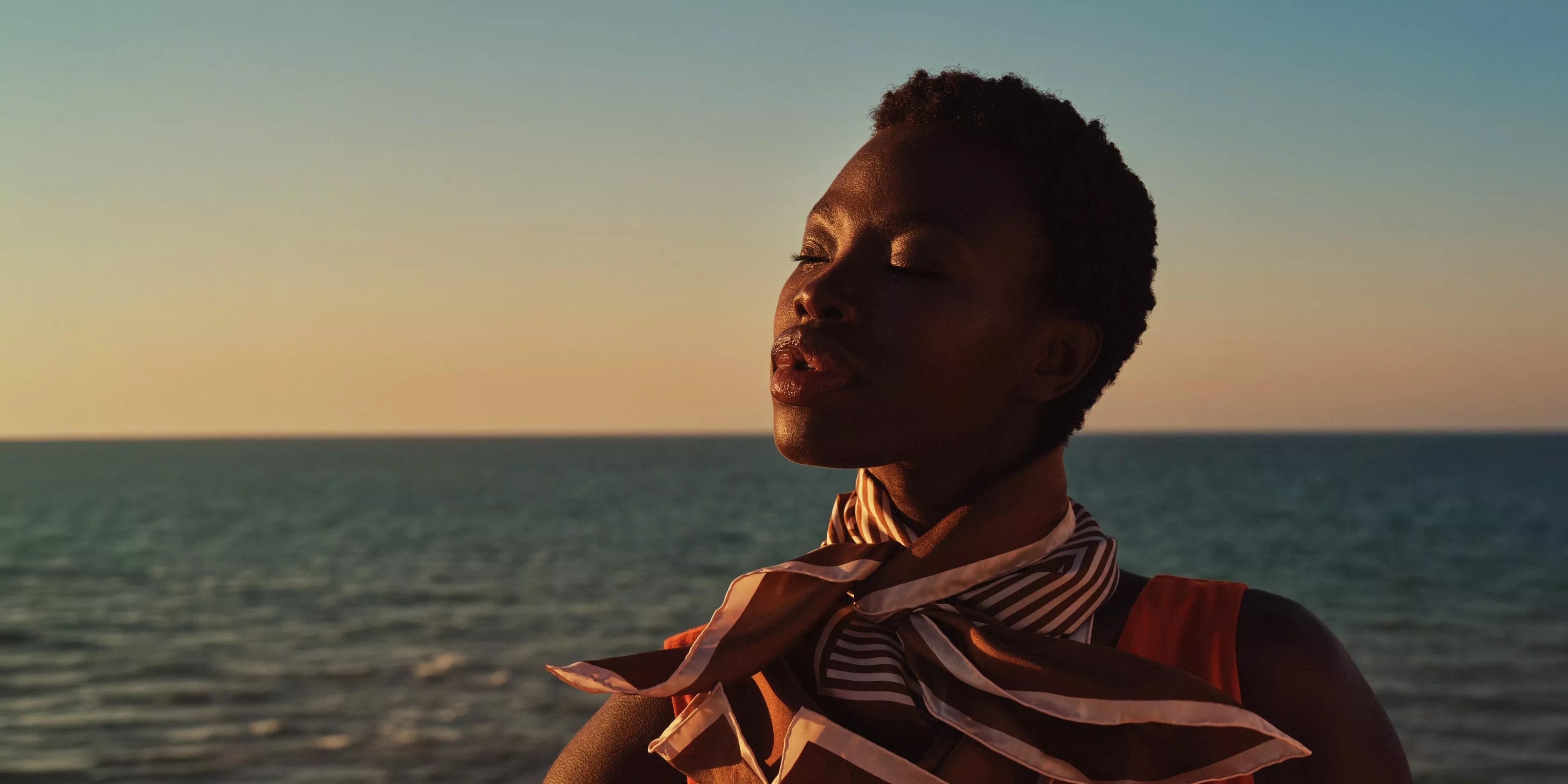 Woman by the sea at sunset, wearing striped scarf and orange top, facing horizon outdoors