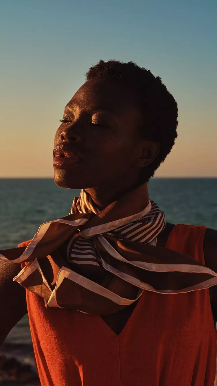 Woman by the sea at sunset, wearing striped scarf and orange top, facing horizon outdoors