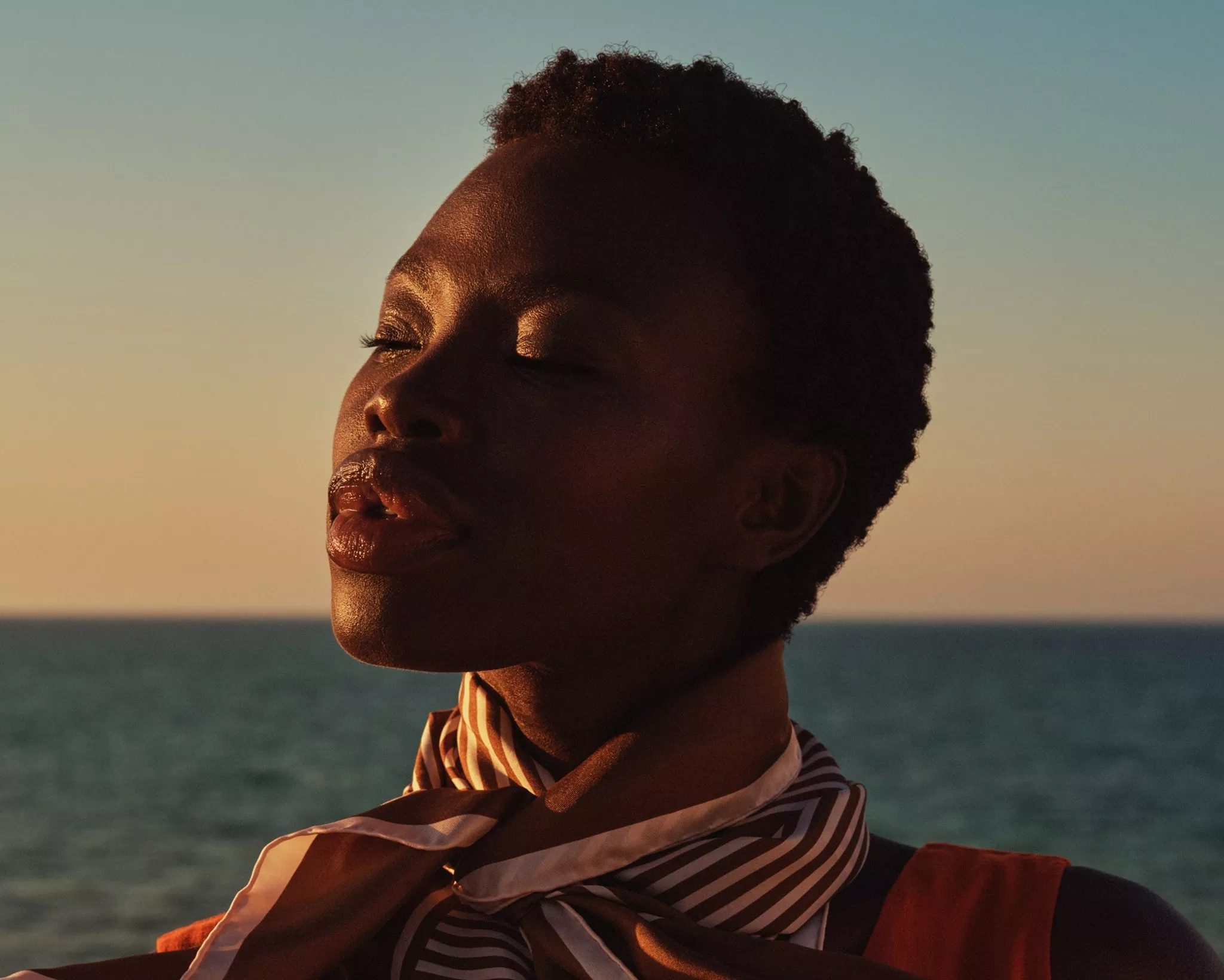 Close-up portrait of a woman by the sea at sunset, eyes closed, wearing a striped scarf in warm light