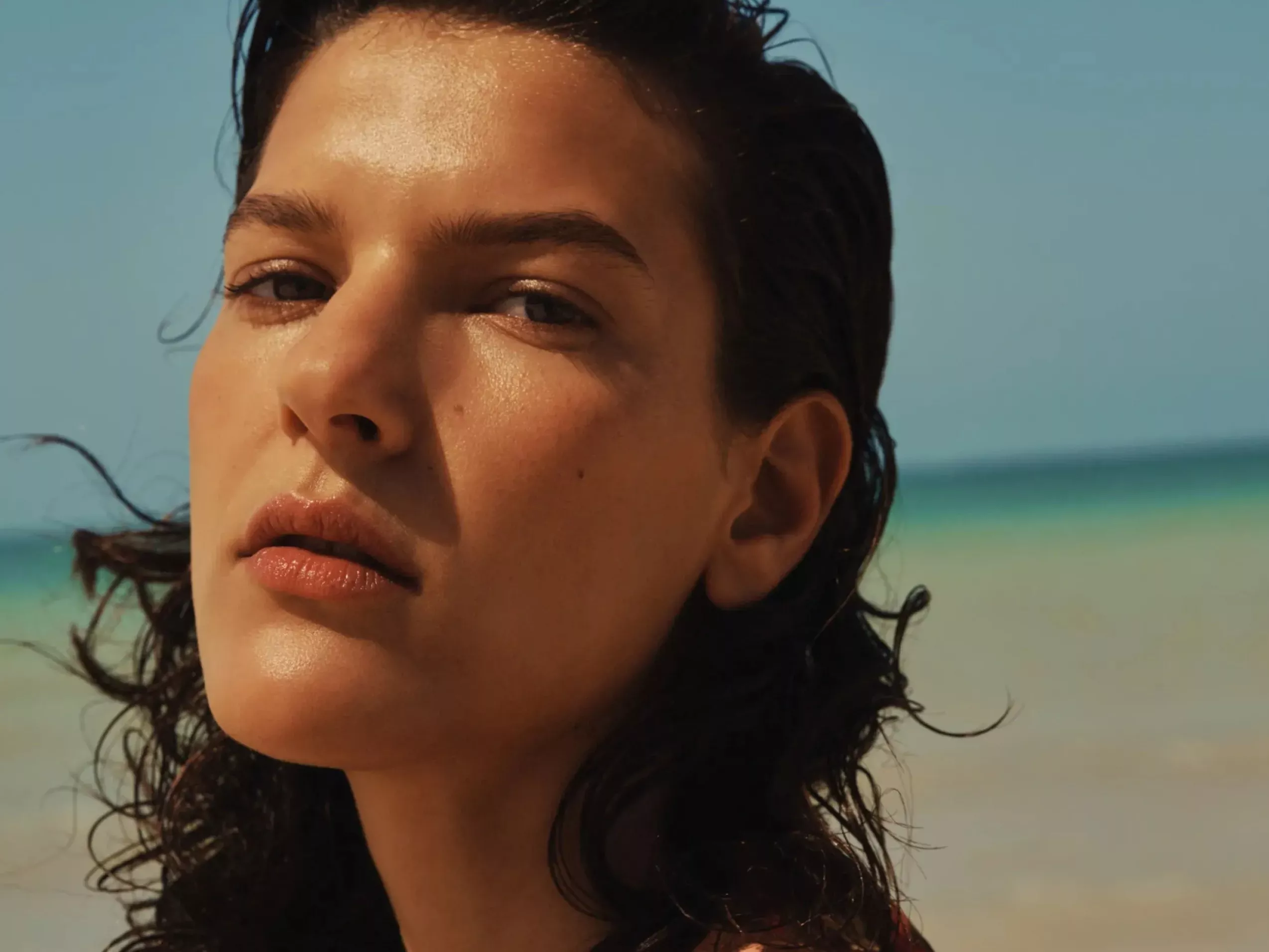 Close-up portrait of a woman on a sunny beach, wet hair and natural makeup, with sea and sky behind.