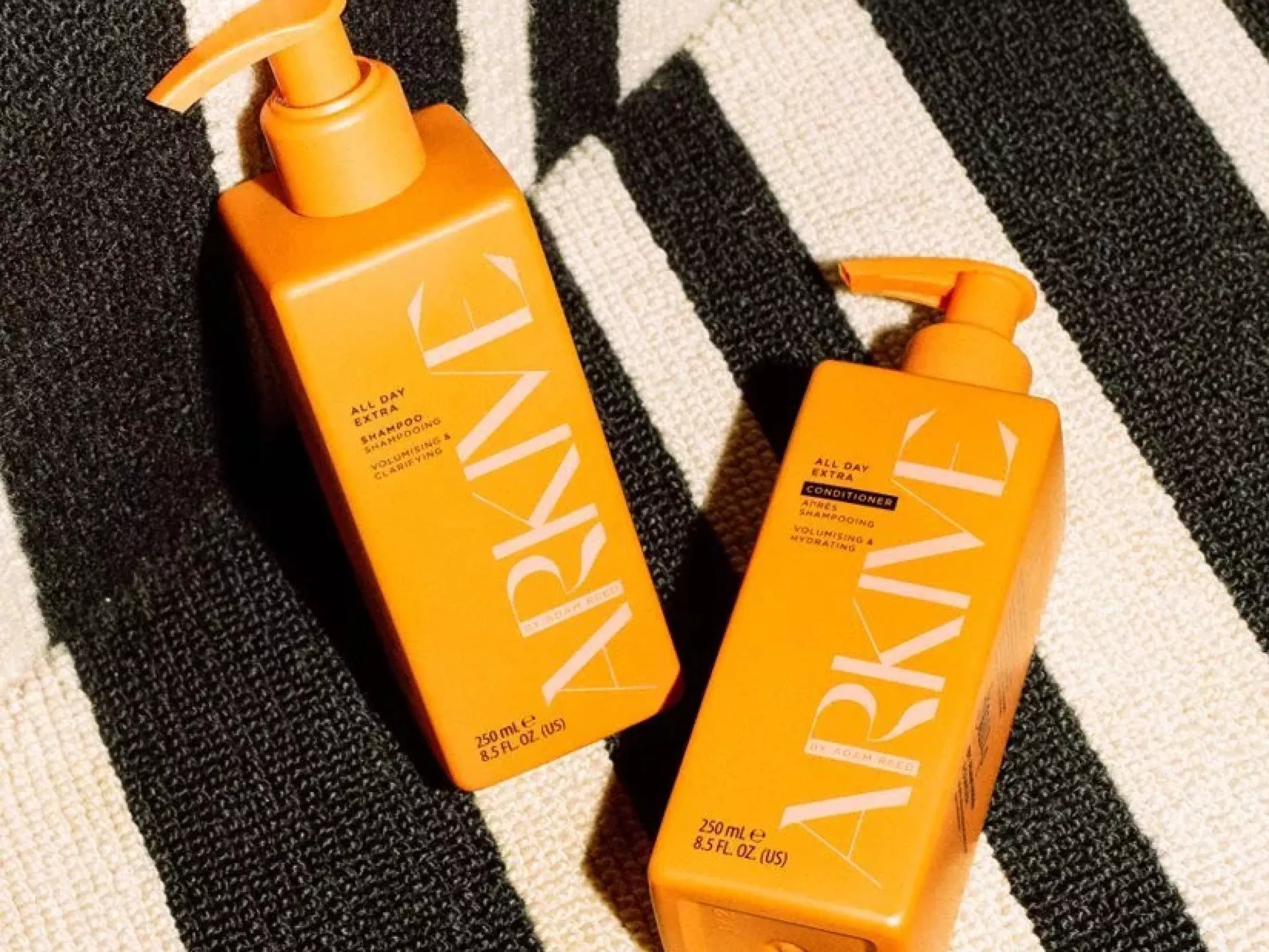 Orange ARKIVE shampoo and conditioner pump bottles on black and white striped towel, summer haircare product shot