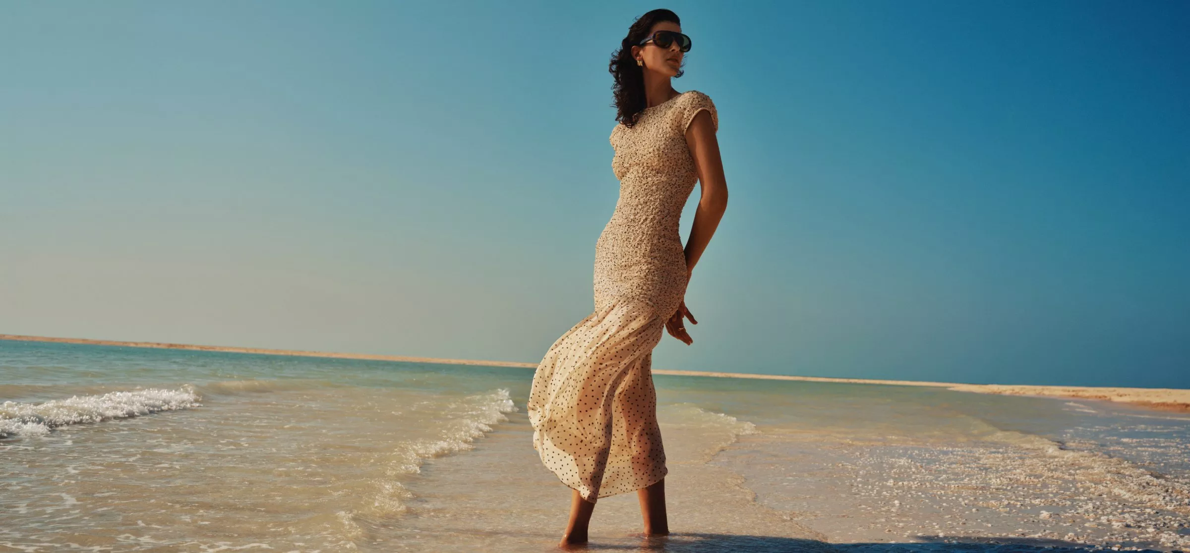 Woman in a cream summer dress standing in shallow sea at sunset, serene beach fashion lifestyle photo