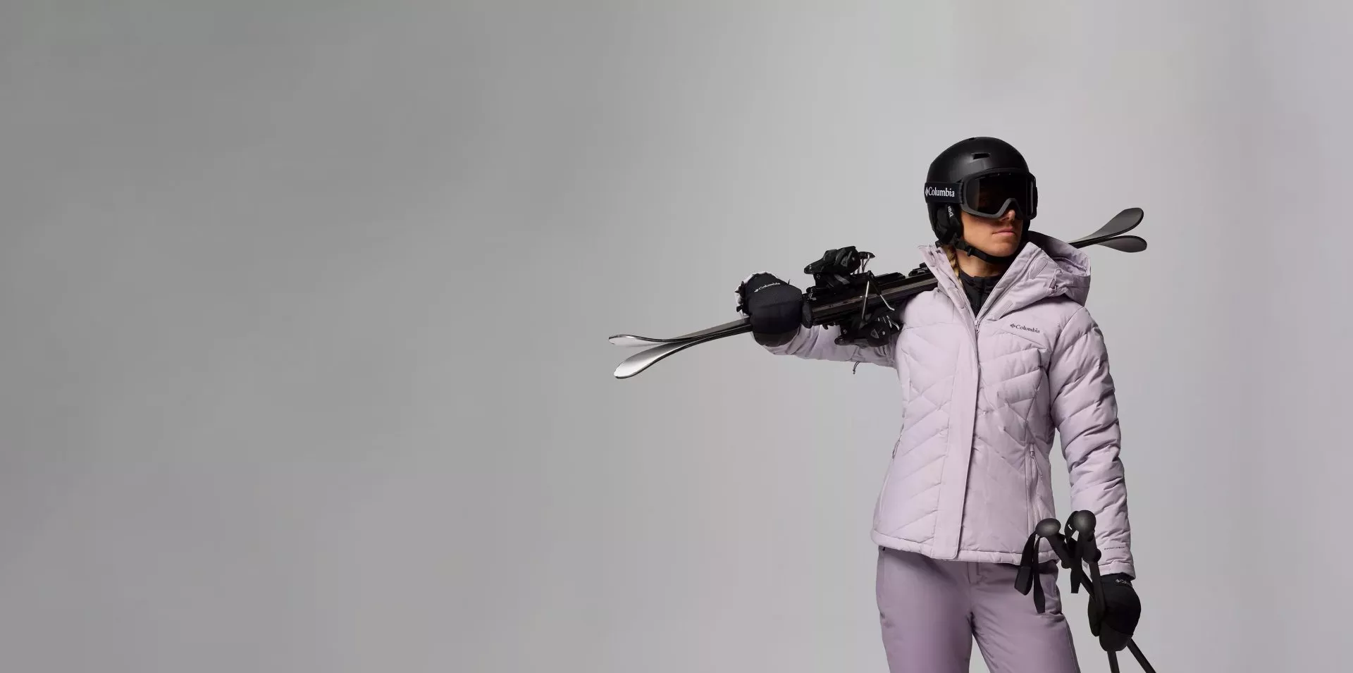 Women in purple ski jacket and ski trousers with ski helmet, goggles and ski equipment with a grey background