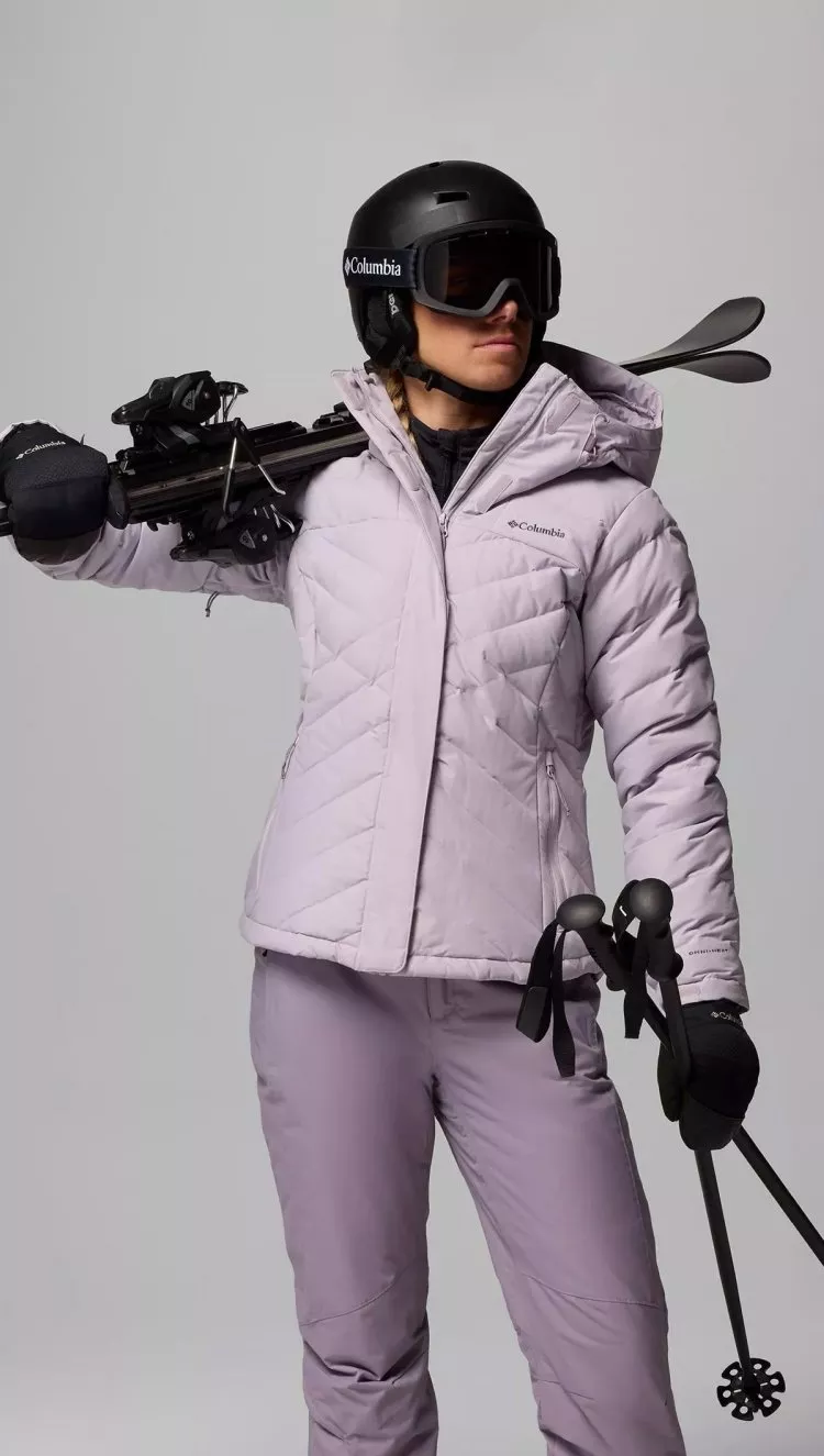 A female model wearing a lilac Columbia ski jacket, holding ski's and wearing a helmet