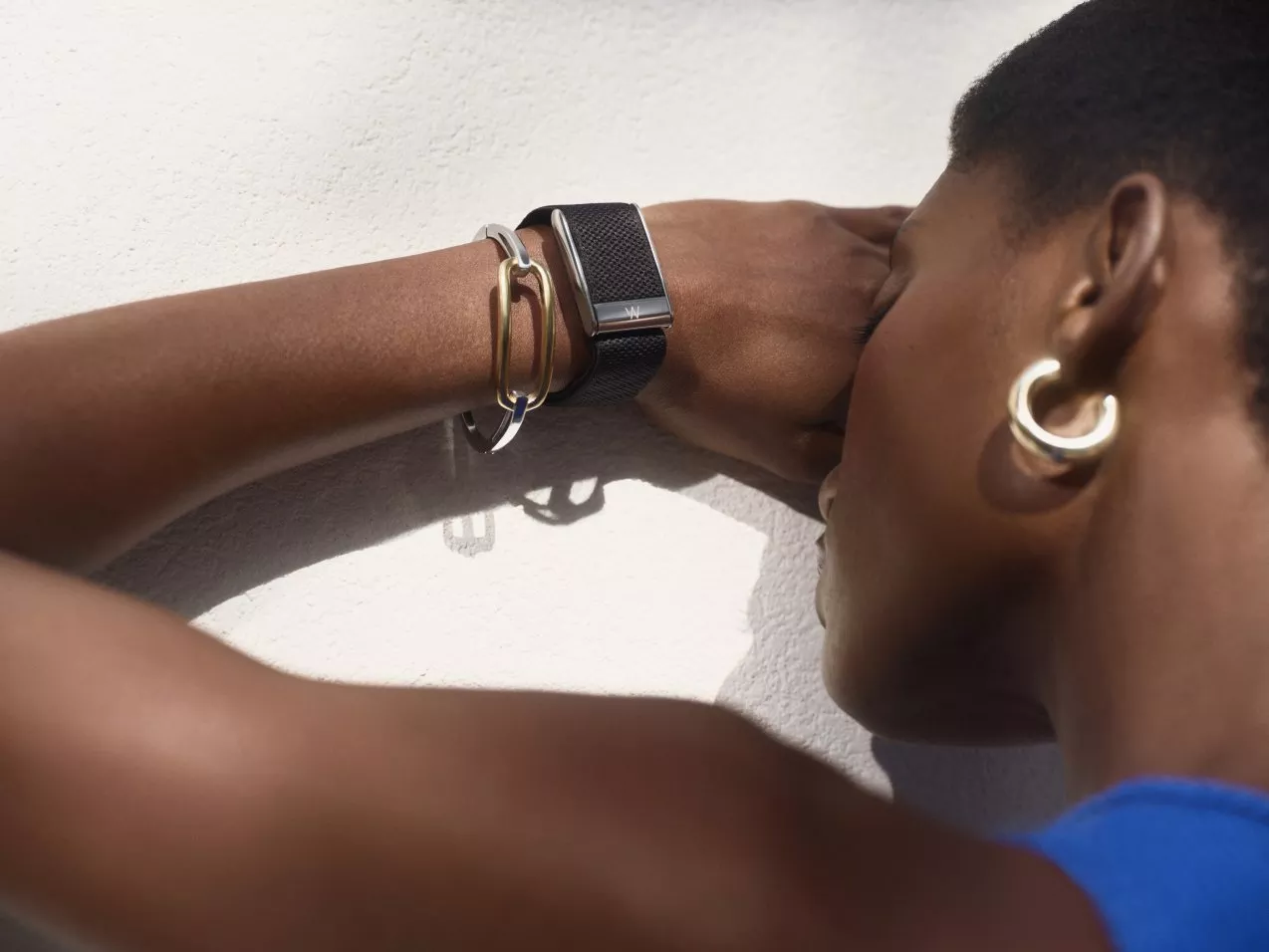 Close-up of woman wearing black mesh watch and gold bangles, leaning on white wall in sunlight