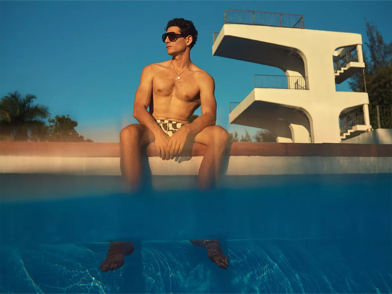Man sitting on the edge of swimming pool in swim shorts, wearing glasses and a chain