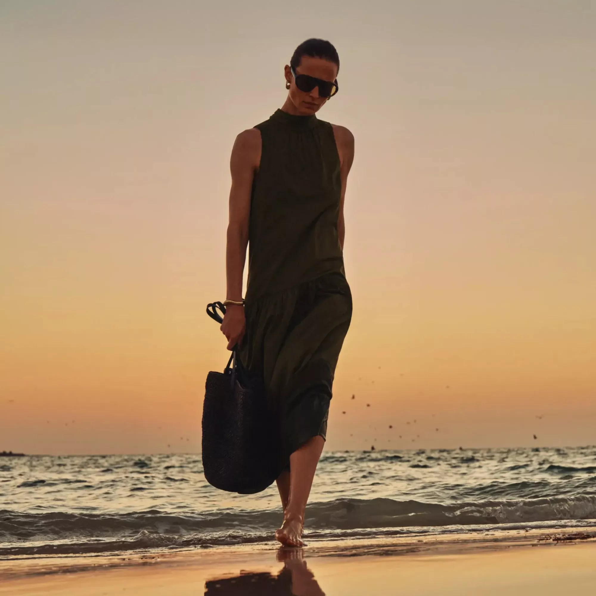 Stylish woman in olive maxi dress and sunglasses walking barefoot on a beach at sunset, holding a tote bag