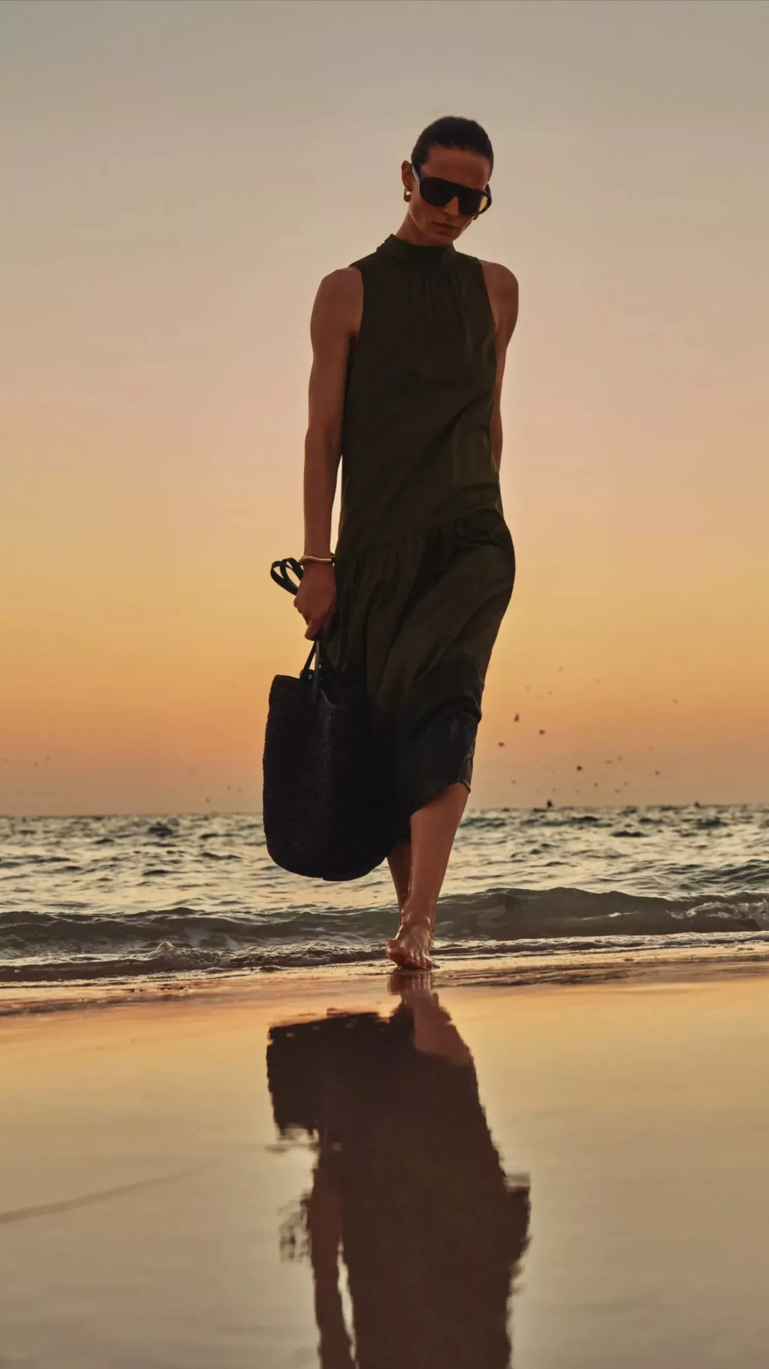 women wearing a dress walking along a beach with a sunset behind her