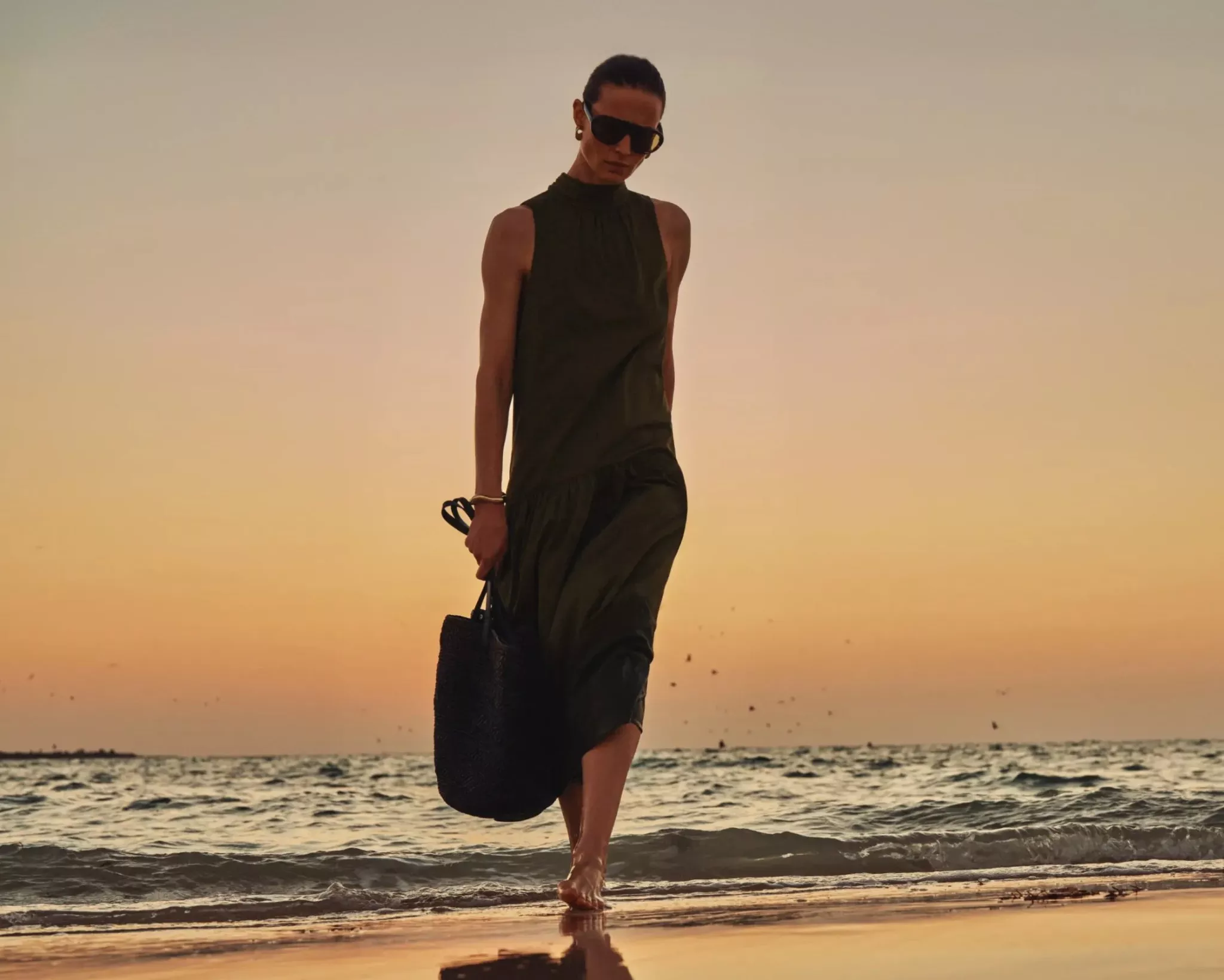 a women walking on a sandy beach in a dress with sunset behind her