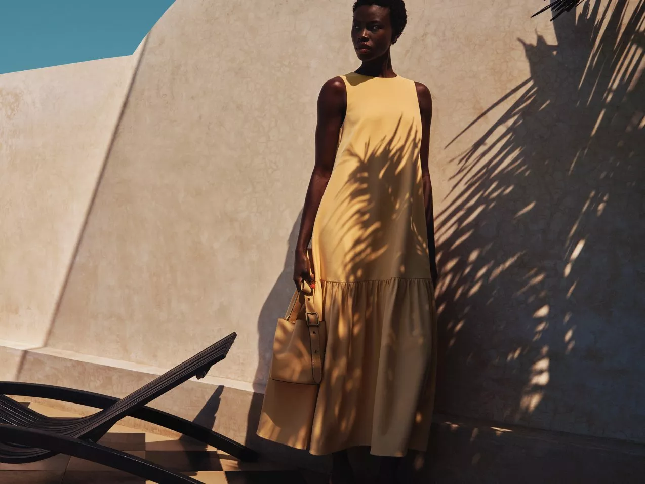 Model in a beige maxi dress holding a leather handbag, standing by a sunlit wall with palm shadows