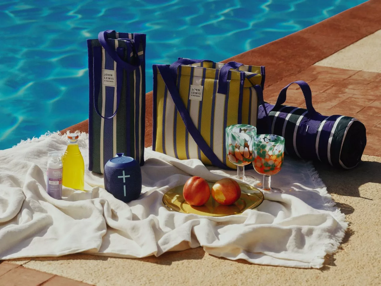 Poolside picnic setup with striped John Lewis bags, blanket, fruit, drinks and patterned glasses by blue water