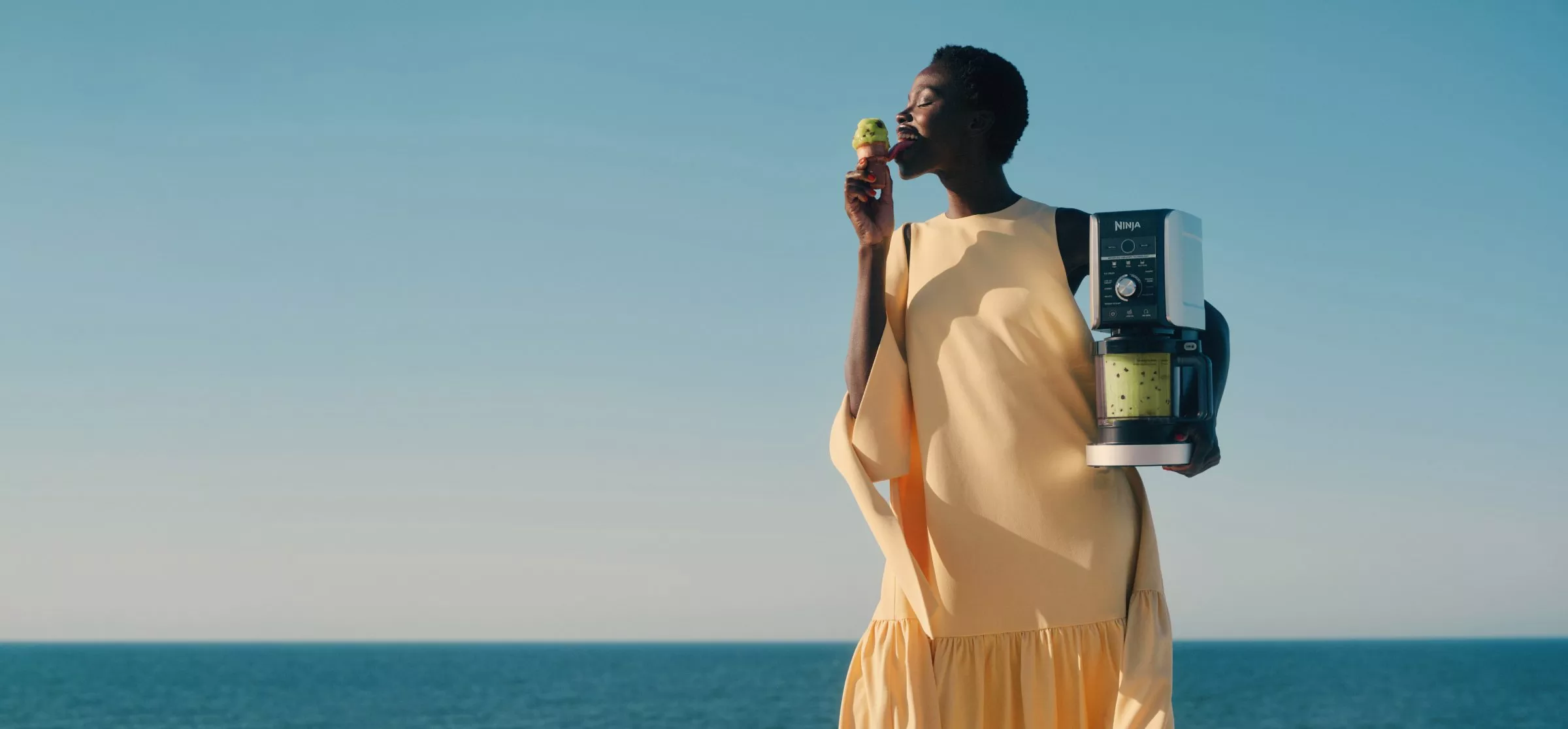 Woman enjoying an ice cream by the sea, holding a Ninja blender with green smoothie in a sunny outdoor scene