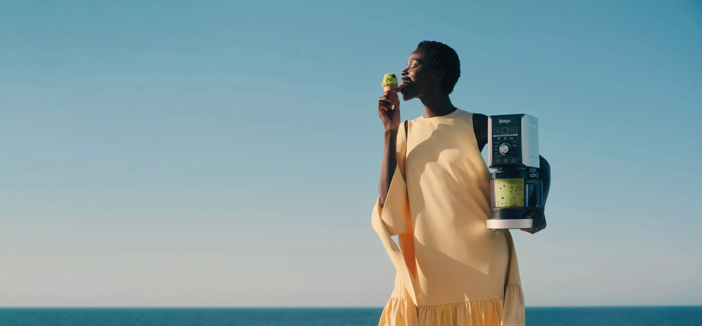 Woman enjoying an ice cream by the sea, holding a Ninja ice cream maker with pistachio gelato in tub