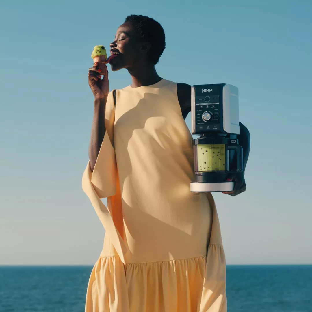 Woman enjoying pistachio ice cream while holding a Ninja blender with green smoothie, outdoors by the sea