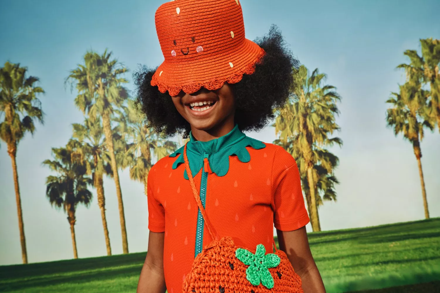 Smiling child in strawberry outfit with orange bucket hat and crochet bag, outdoors with palm trees and blue sky