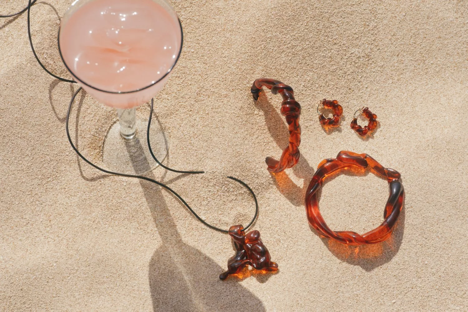 Pink cocktail in a glass on sand with tortoiseshell resin jewellery set, bracelet, earrings and pendant necklace