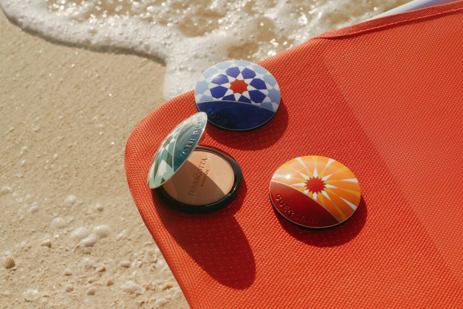 Guerlain Terracotta bronzer compacts on a red beach towel beside sea foam, summer makeup product flat lay.
