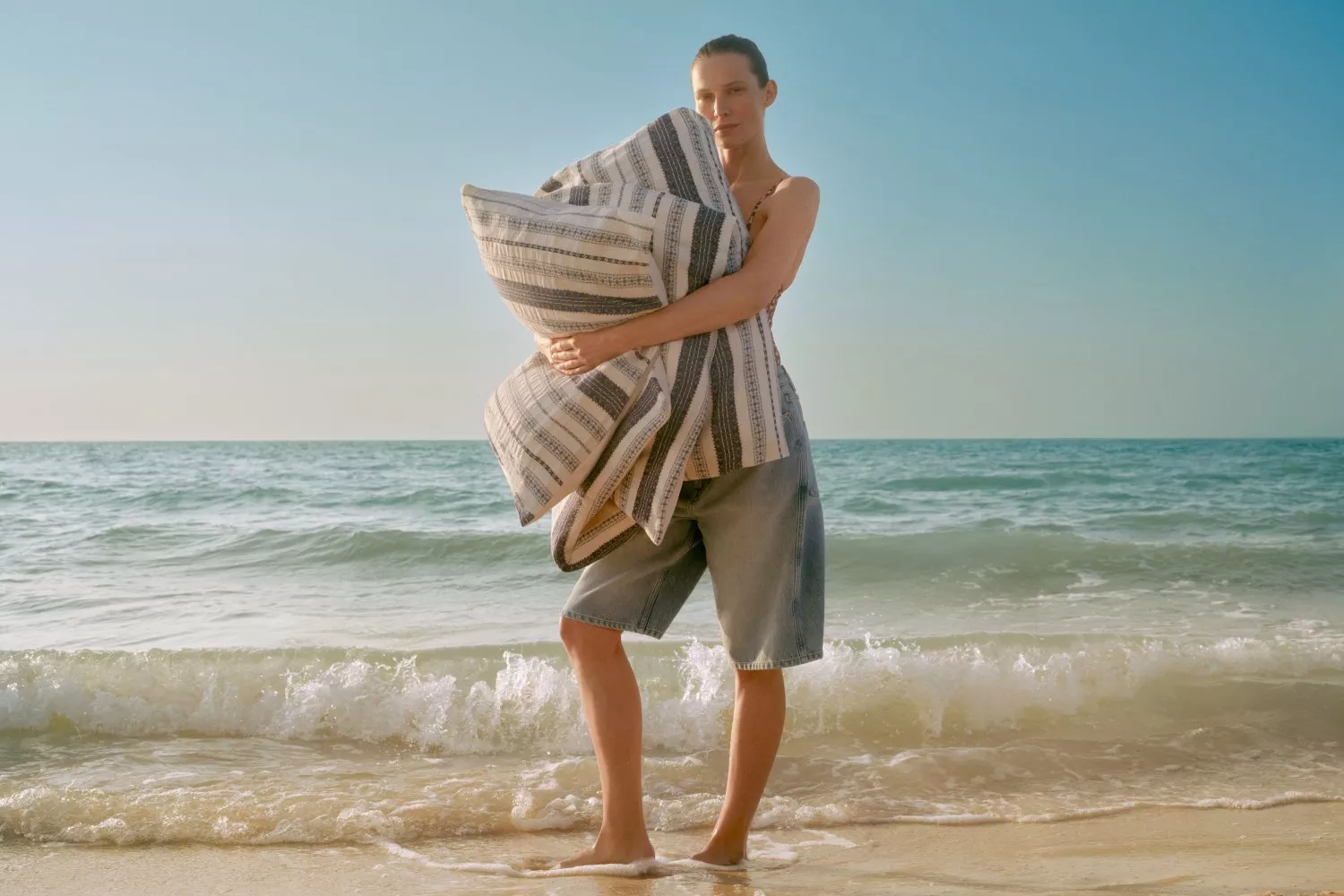 Woman standing on sandy beach holding a striped duvet, summer coastal lifestyle bedding photo by the sea