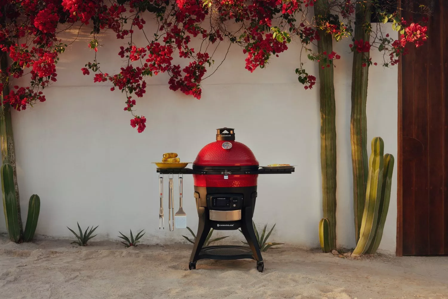 Red ceramic kamado barbecue grill outdoors by white wall, cacti and red bougainvillea, with tools and corn.