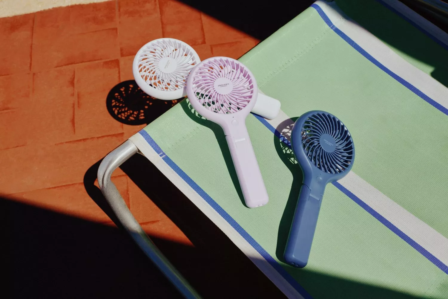 Three portable handheld mini fans on a green striped sun lounger, outdoor summer cooling accessories in sunlight