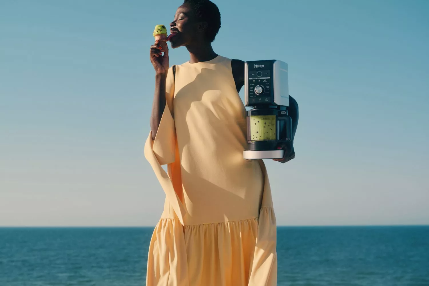 Woman holding Ninja ice cream maker by the sea, licking a scoop of pistachio gelato under blue sky