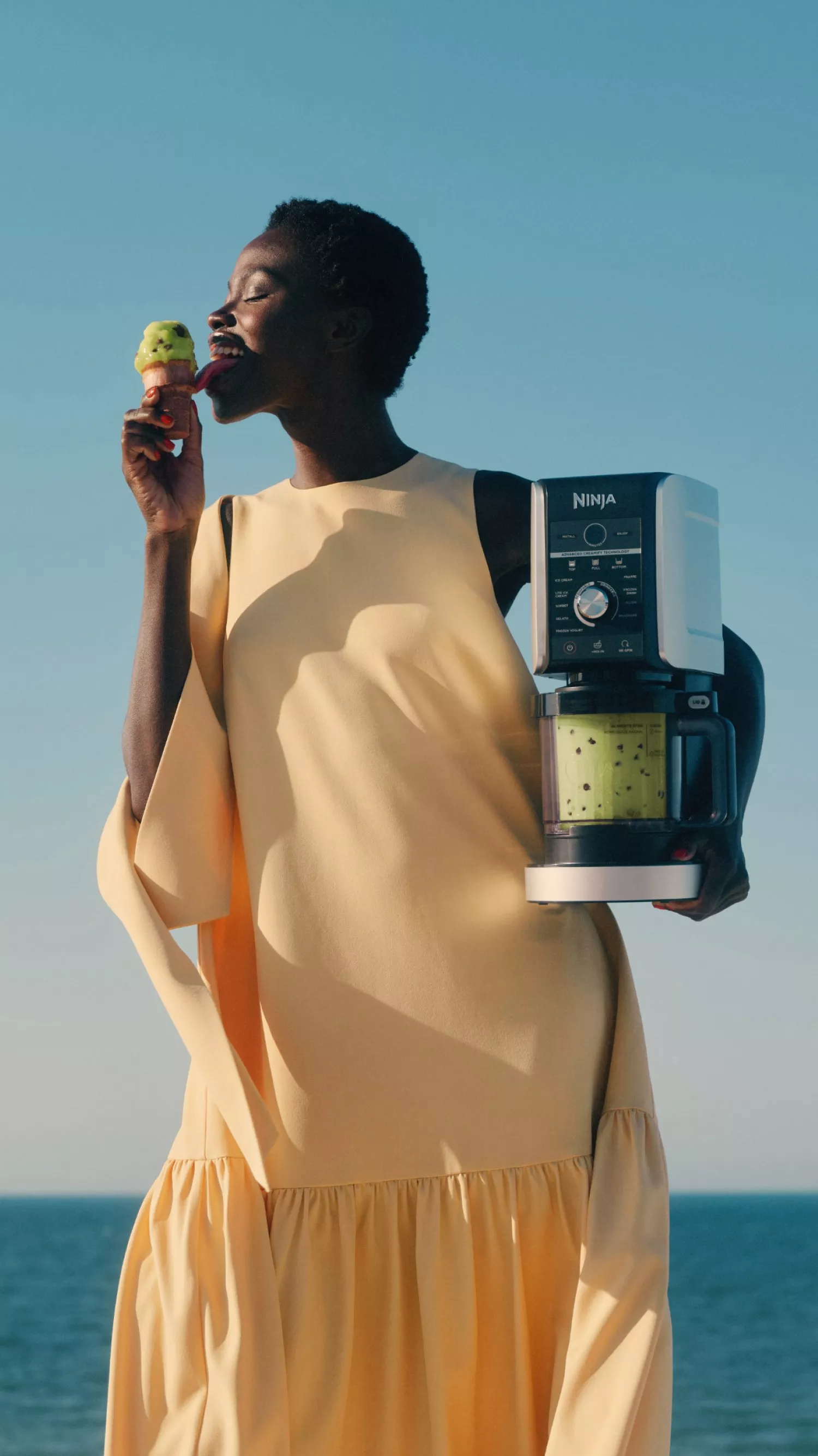 Woman eating green ice cream while holding Ninja blender, seaside background, summer lifestyle product photo