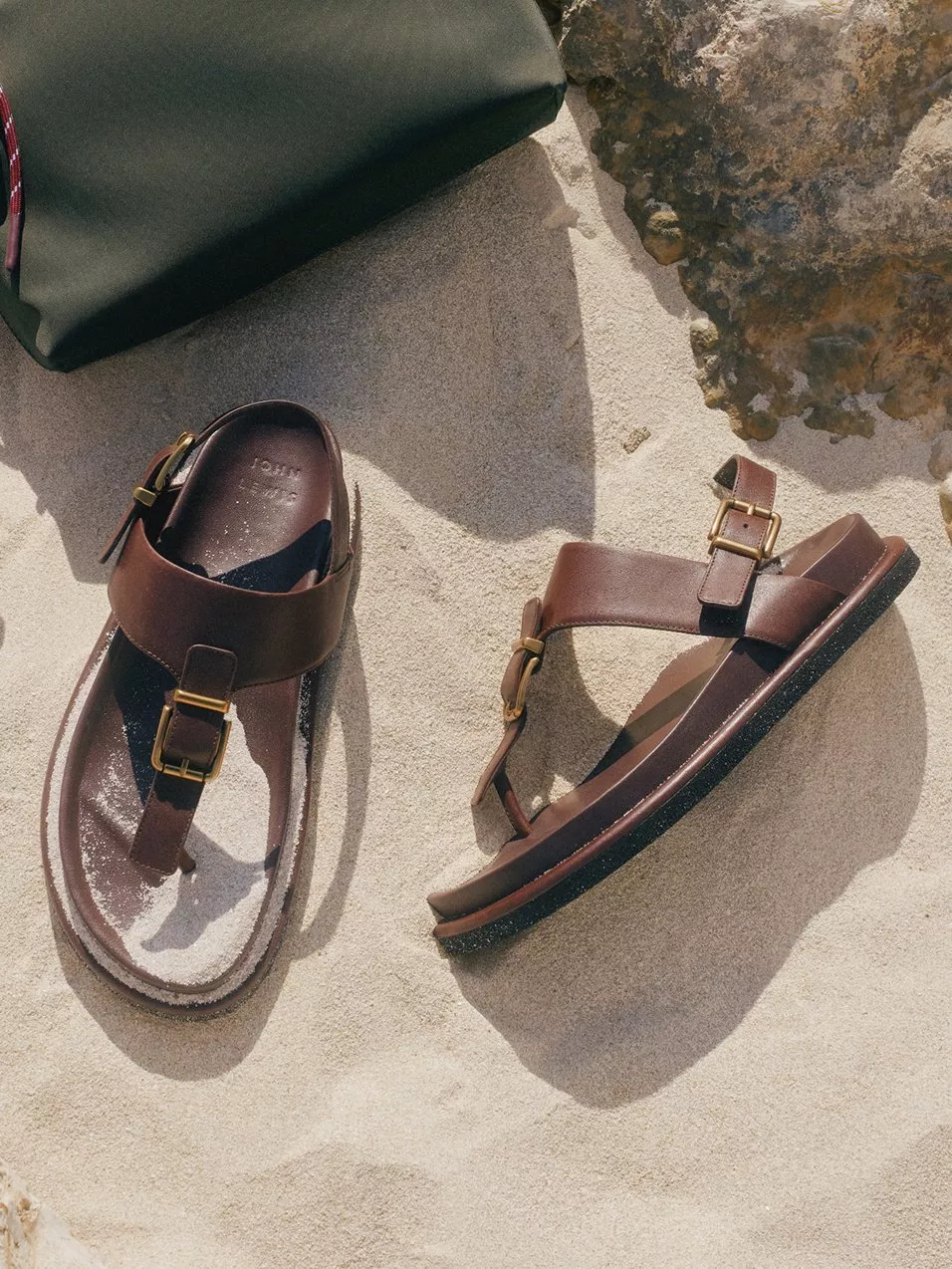 Brown leather sandals and a green crossbody bag on sandy beach rocks