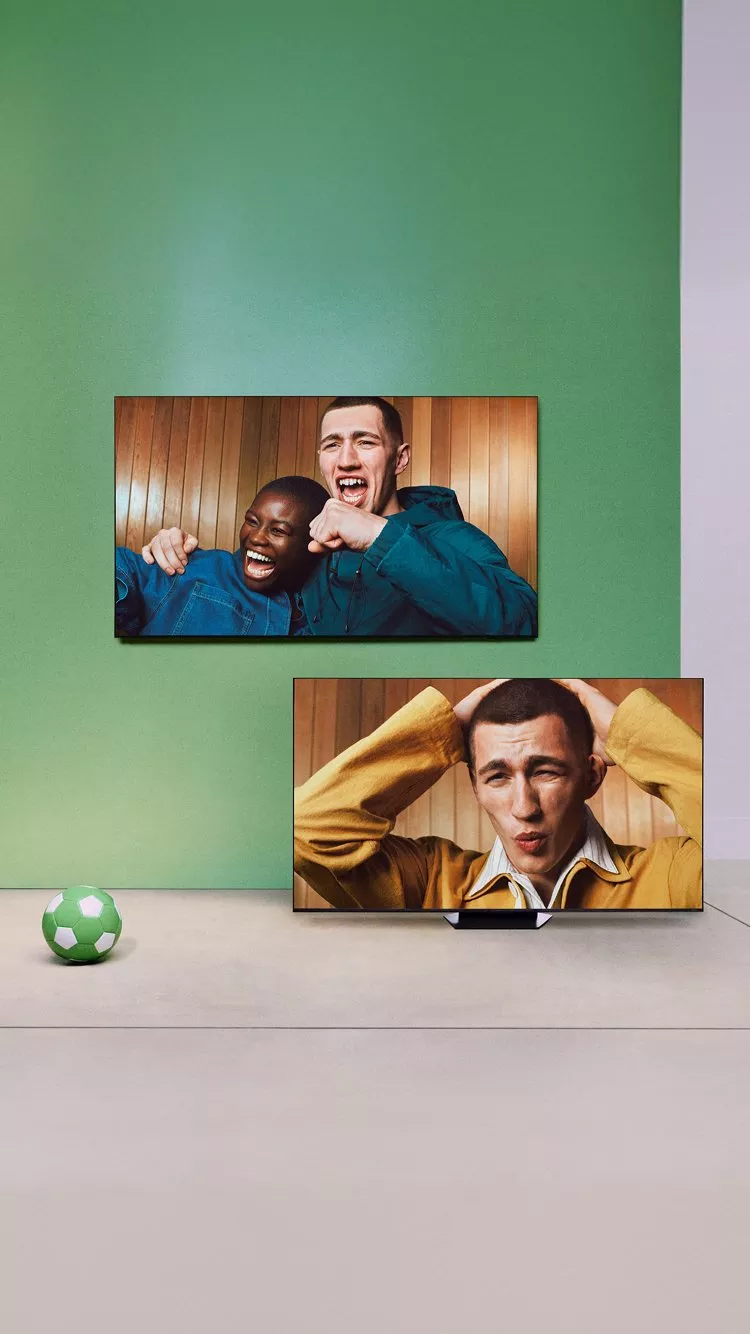 Two TVs show football fans cheering and reacting, with a green football on the floor in a minimalist room.