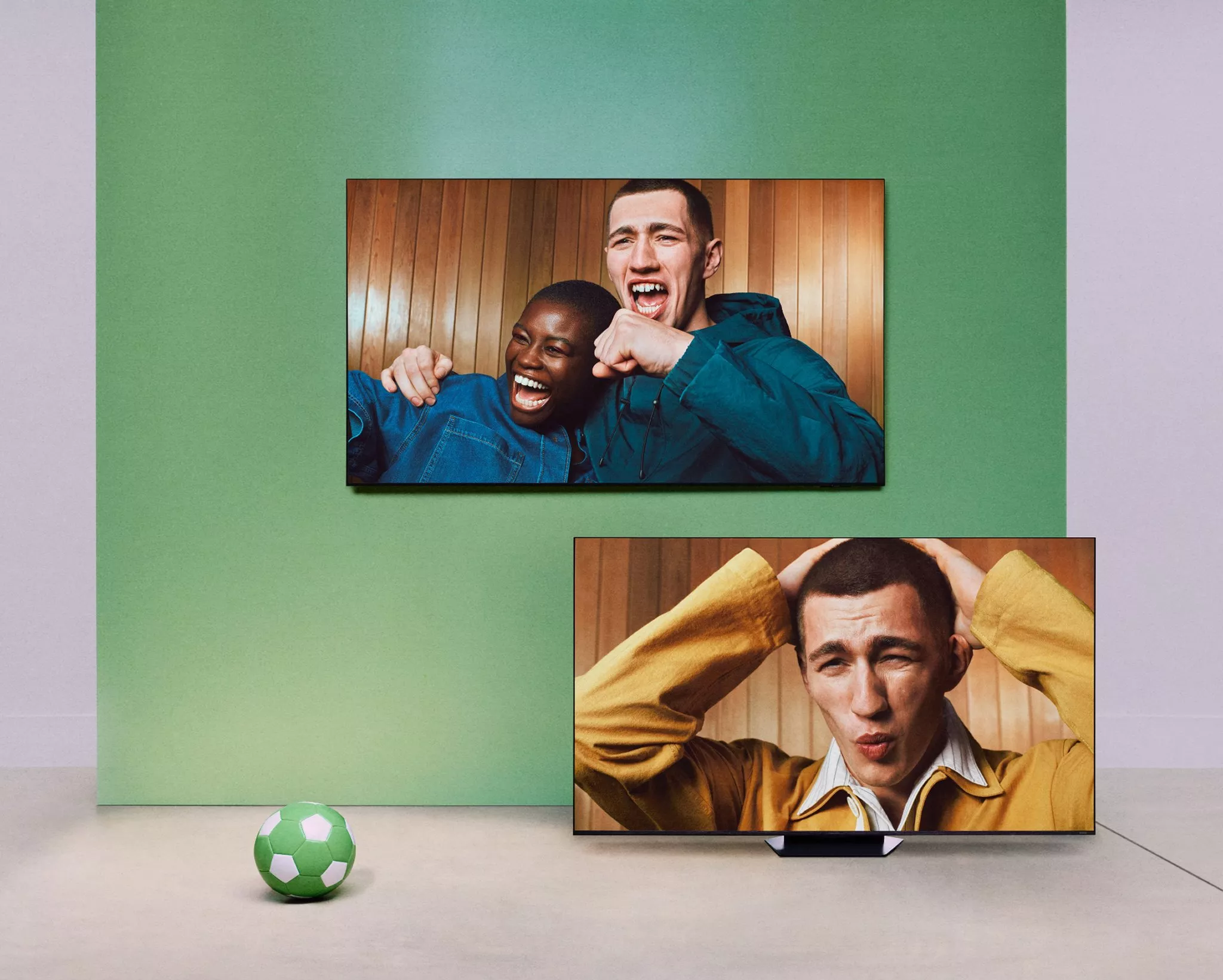 Two TVs show football fans cheering and reacting, with a green football on the floor in a modern room.
