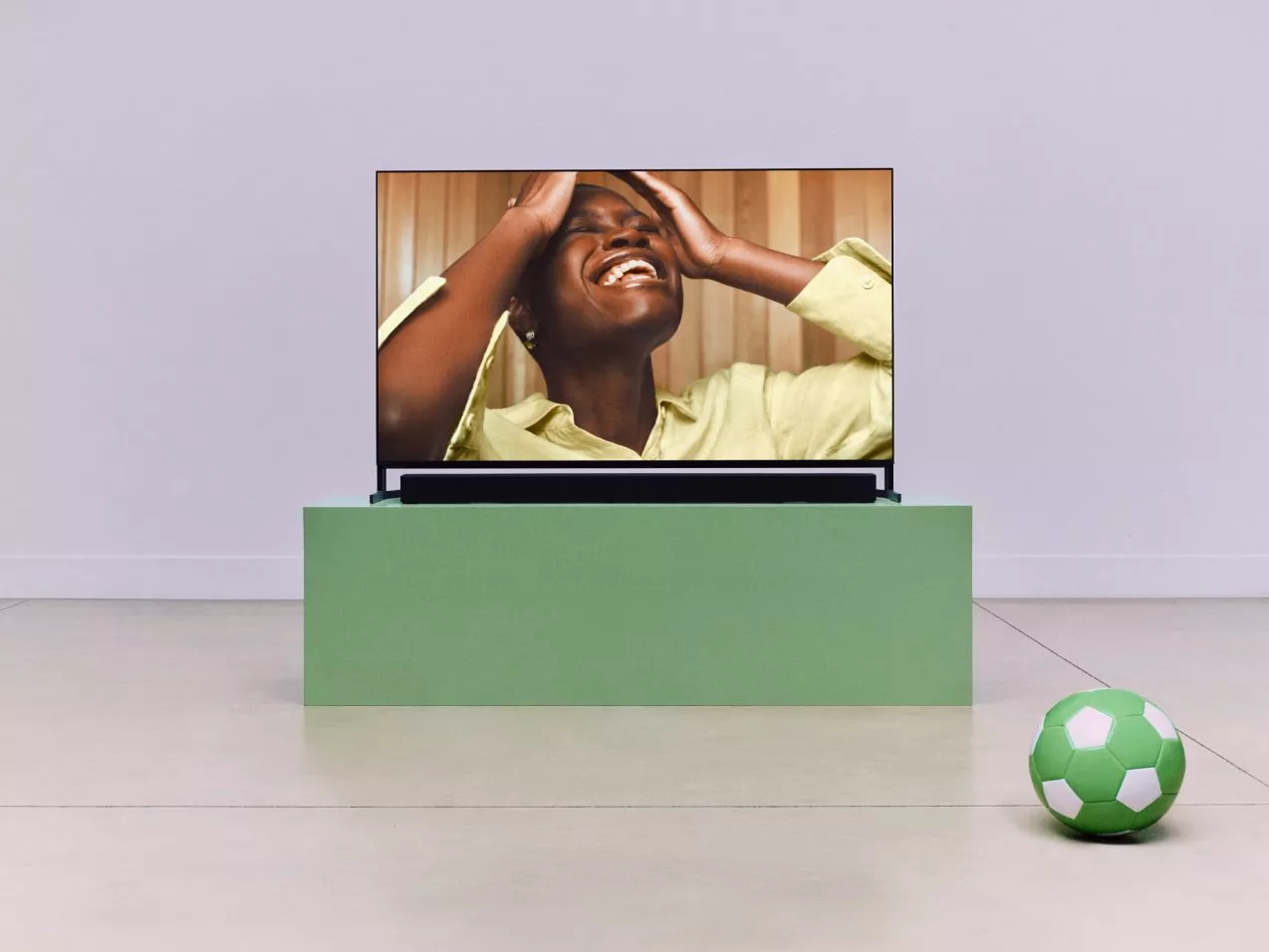 Sony Bravia 8 II TV on green stand showing a woman laughing, with a green football on the floor nearby