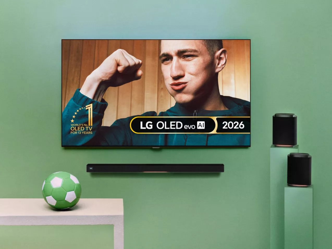 Wall-mounted LG W6 OLED Wallpaper TV showing a man flexing, with soundbar, speakers and green football nearby
