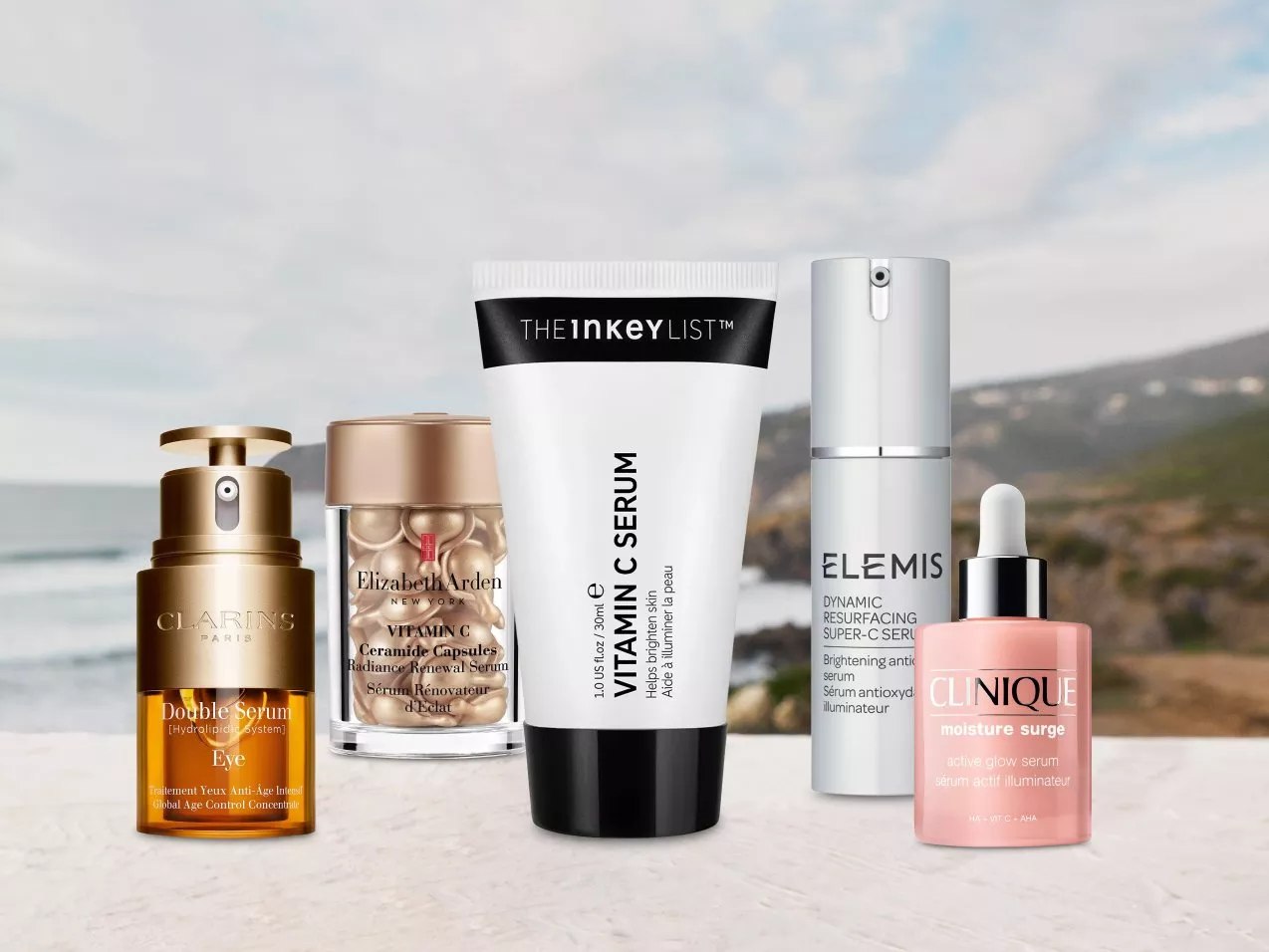 A selection of skincare serums from various brands displayed in front of a scenic coastal background.