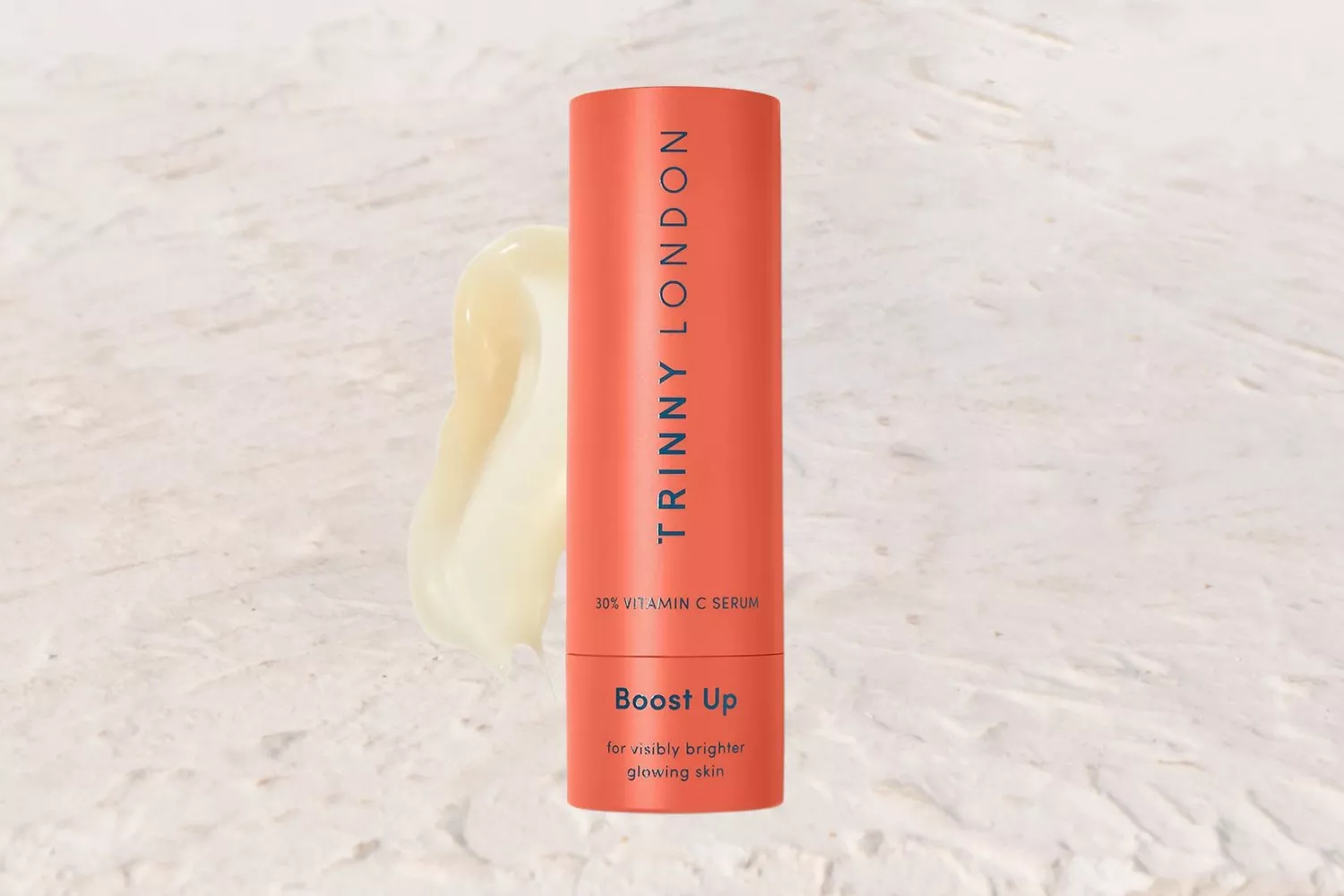 Orange Trinny London Vitamin C serum bottle on a light textured background with a smear of cream beside it.