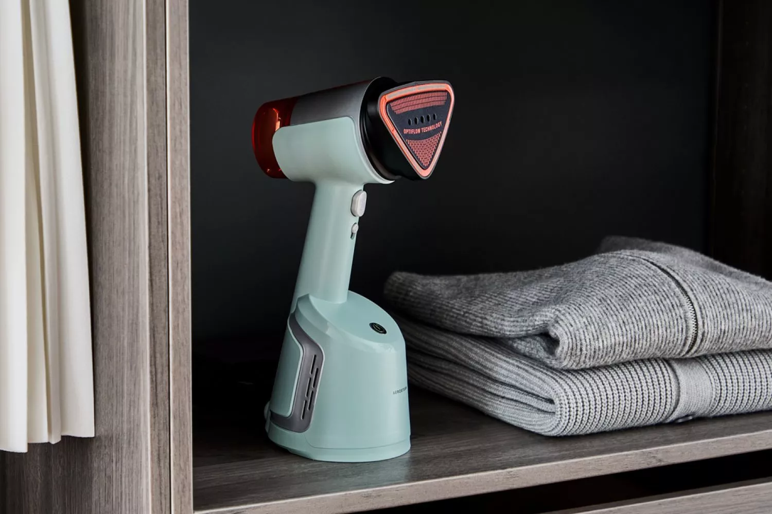 Handheld garment steamer on wardrobe shelf beside folded knit jumpers, home laundry and clothing care device