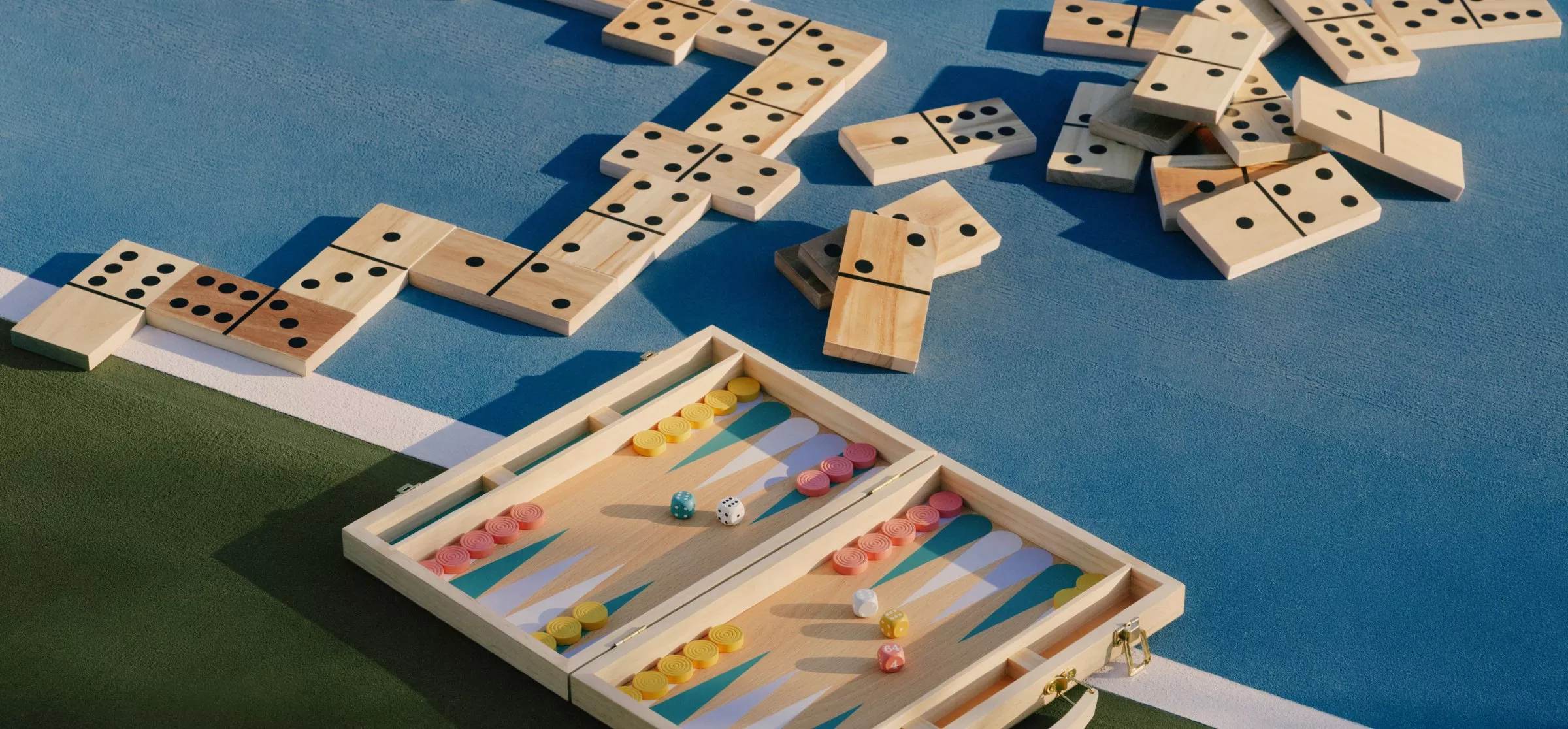 Wooden backgammon set with dice and counters beside scattered domino tiles on a blue outdoor court surface