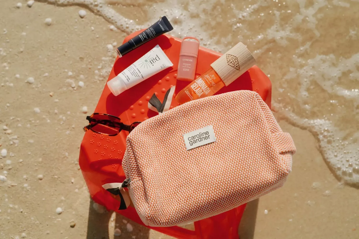 Beach skincare essentials on sand: Caroline Gardner wash bag, sunglasses, sunscreen and lip balm on orange mat