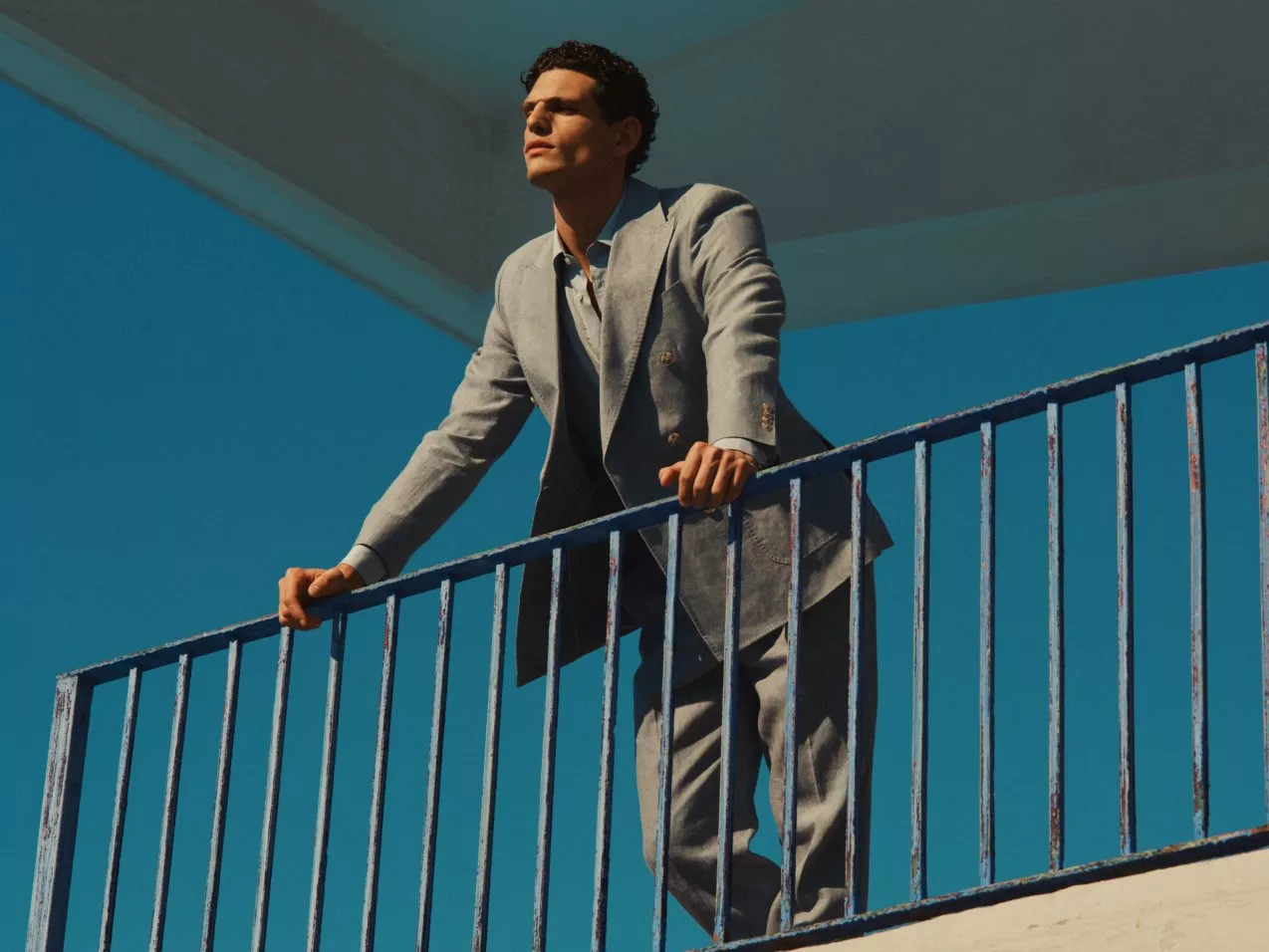 Stylish man in a light grey suit leaning on balcony railing, sunlit modern architecture and blue sky backdrop