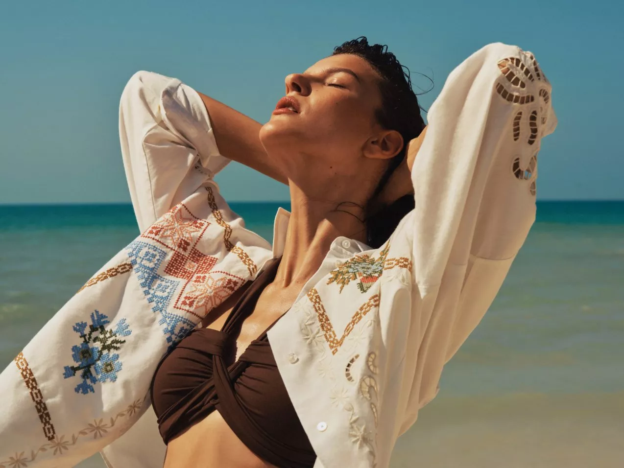 Woman in embroidered white shirt and brown bikini top posing by the sea on a sunny beach holiday