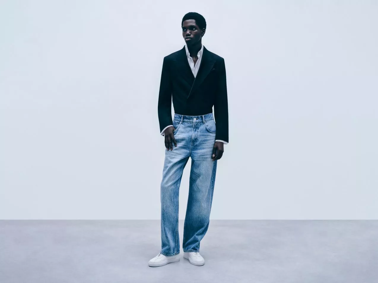 Male fashion model in black blazer and white shirt with light blue jeans and trainers on studio backdrop
