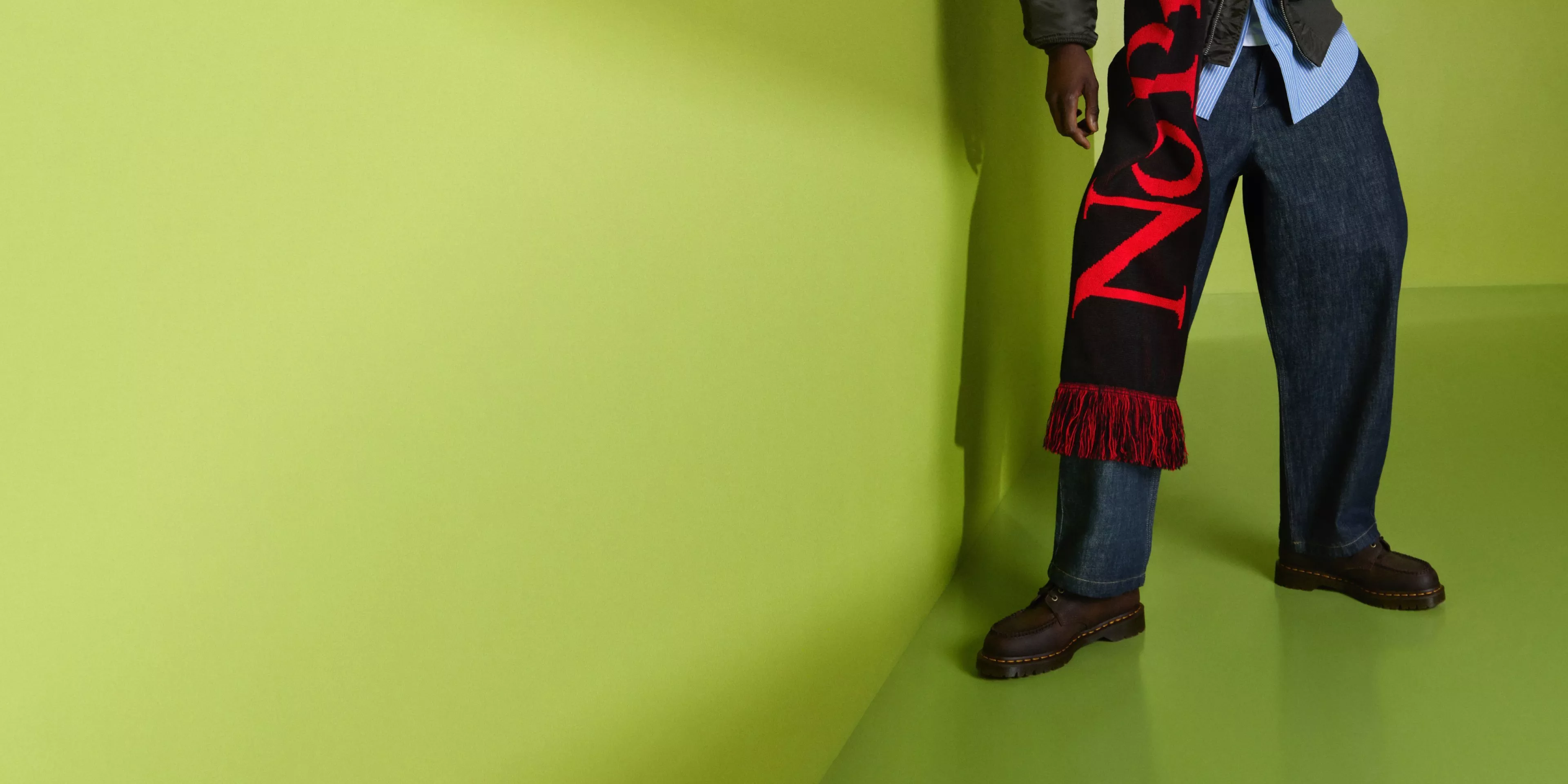 Fashion model in jeans holding black scarf with red lettering and fringe against a bright green studio backdrop