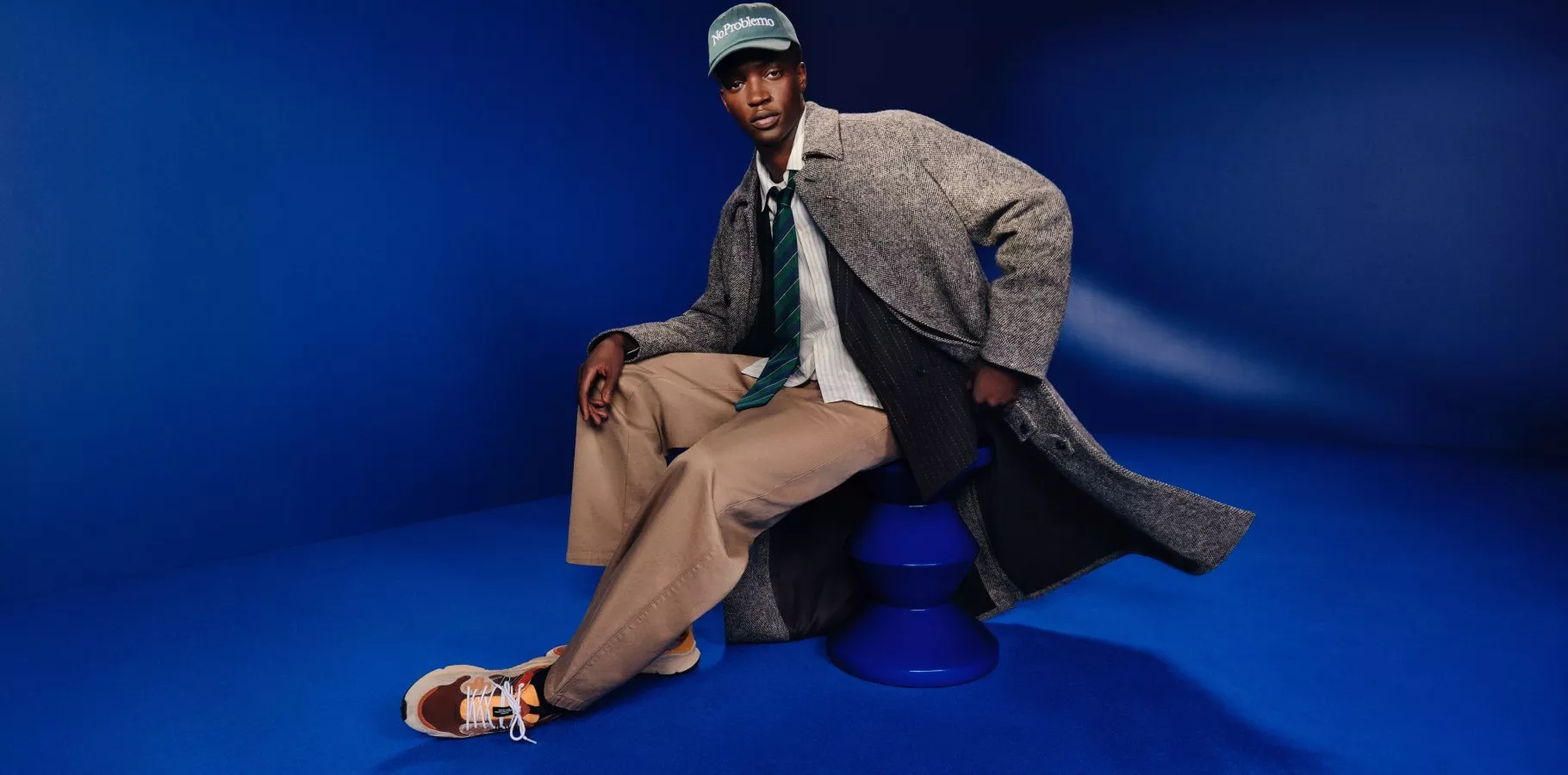 Menswear fashion portrait of a man seated on a blue stool, wearing a grey overcoat, tie and cap on blue backdrop