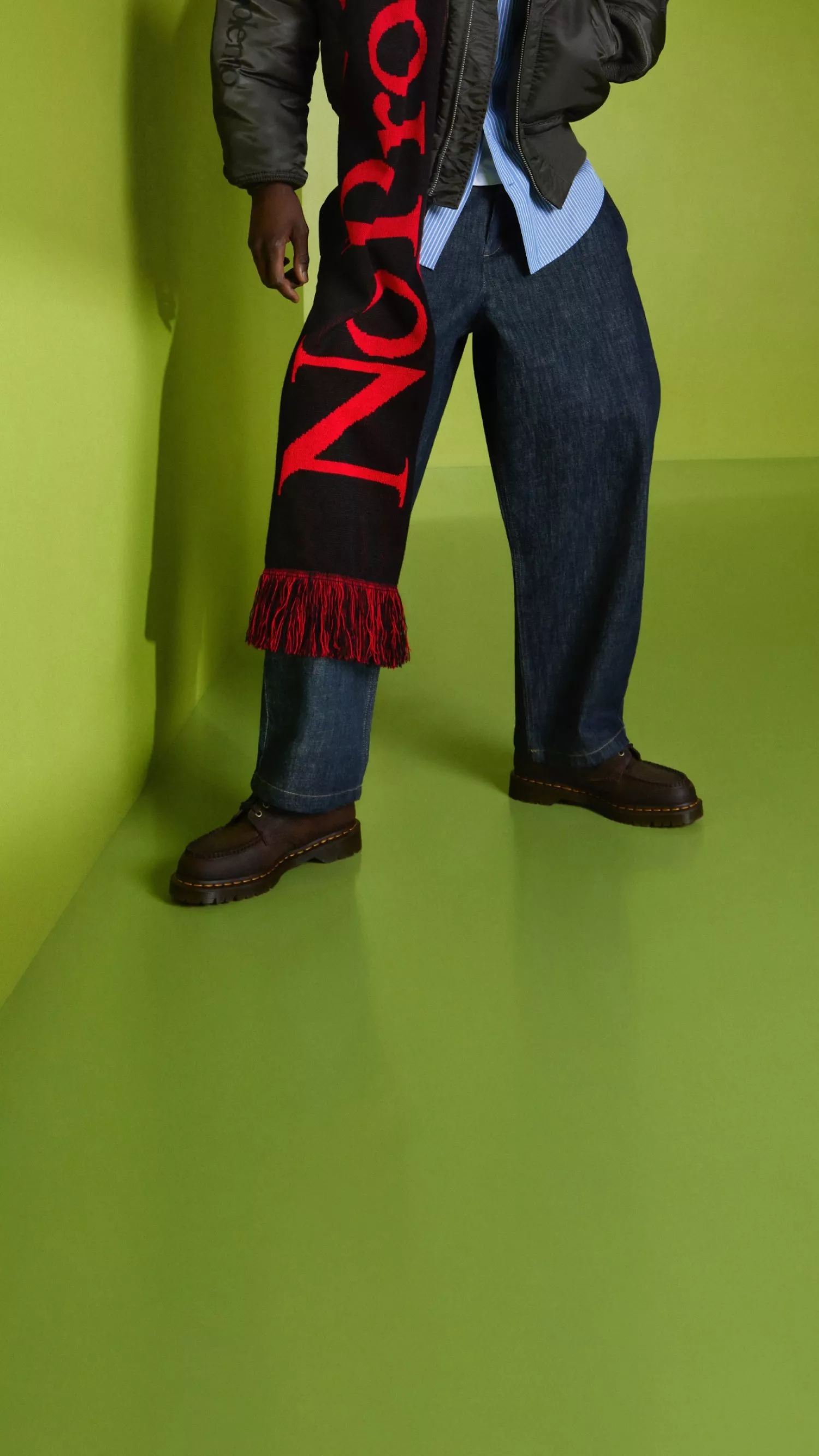 Model in blue jeans and black leather boots with a bold red fringed logo scarf, studio fashion shot