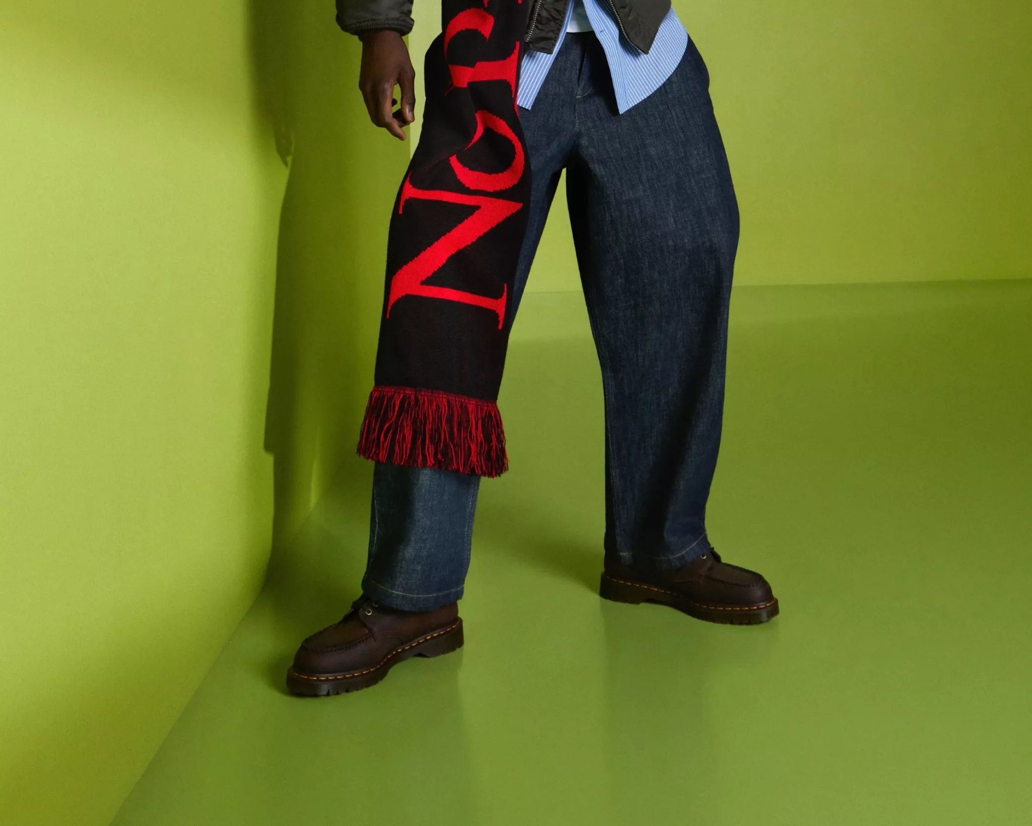 Fashion model wearing dark blue jeans and brown leather boots, with a black red fringed logo scarf on green backdrop